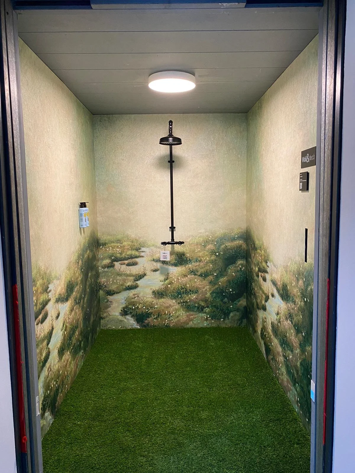 Interno di una cabina con pareti decorate come un paesaggio naturale, pavimento in erba sintetica, e una doccia al centro sotto una luce circolare.