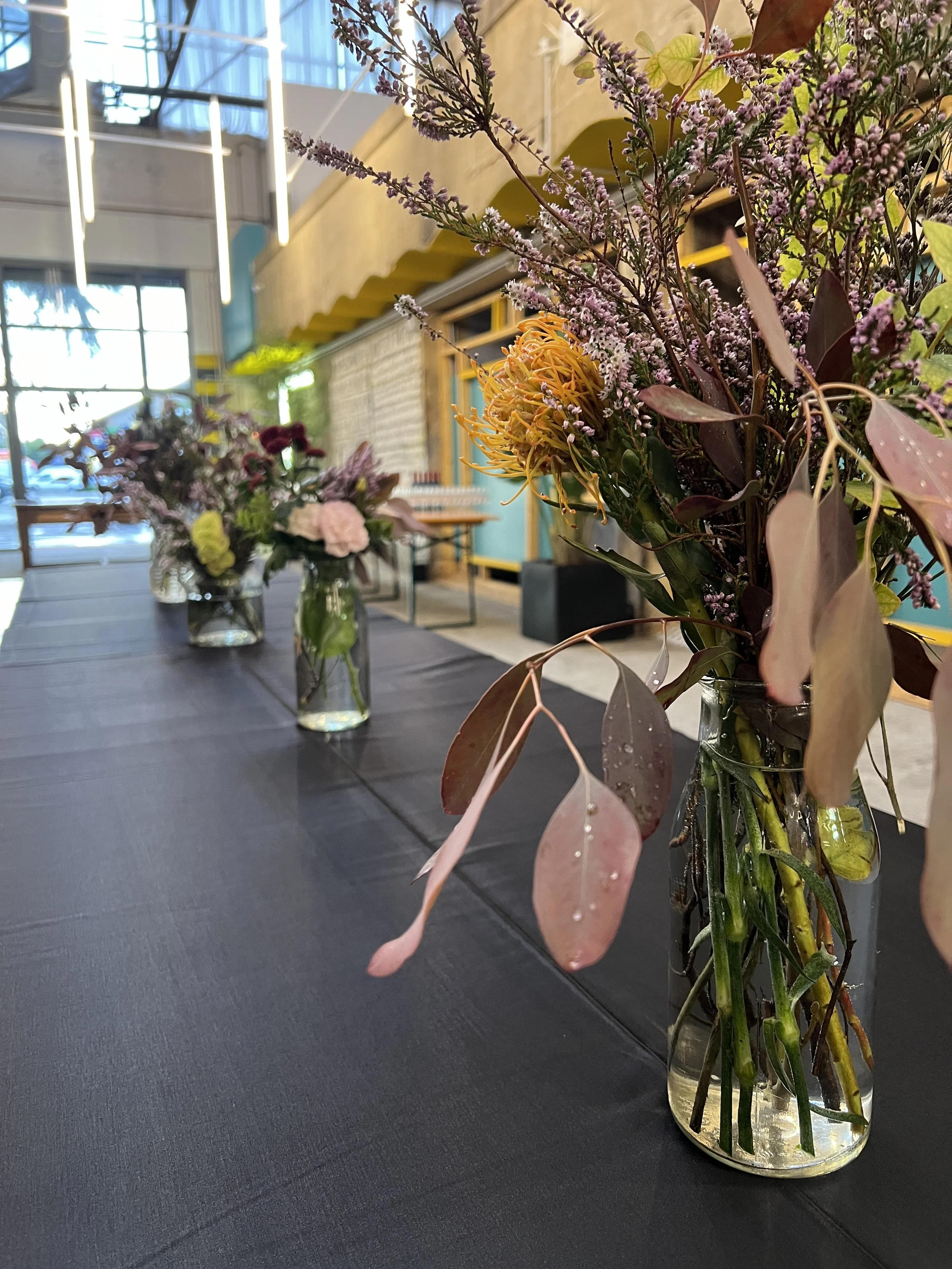 Vasi di vetro con fiori colorati su un tavolo coperto da una tovaglia nera in un ambiente interno con illuminazione moderna.