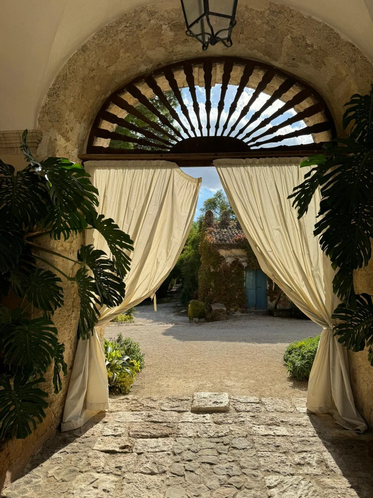 Ingresso ad un cortile di una commenda in Sicilia, con portico ad arco in pietra, tende bianche aperte e piante tropicali sui lati.
