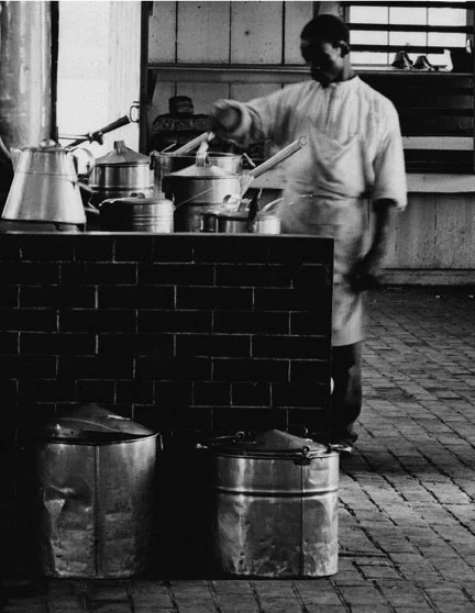 This cook has his hands full with numerous pots and kettles on an impressive stove. Notice the variety of lids and handles. Also note the bails ears and handles of the large kettle on the floor. These vary from the simplified style seen in most camps.