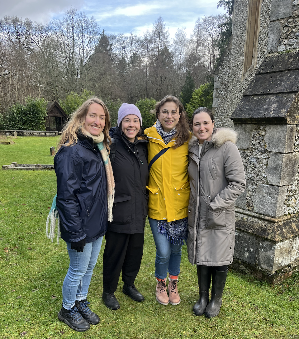 Four women wearing coats stand smiling outside of a church with sparse winter woodland behind in the background