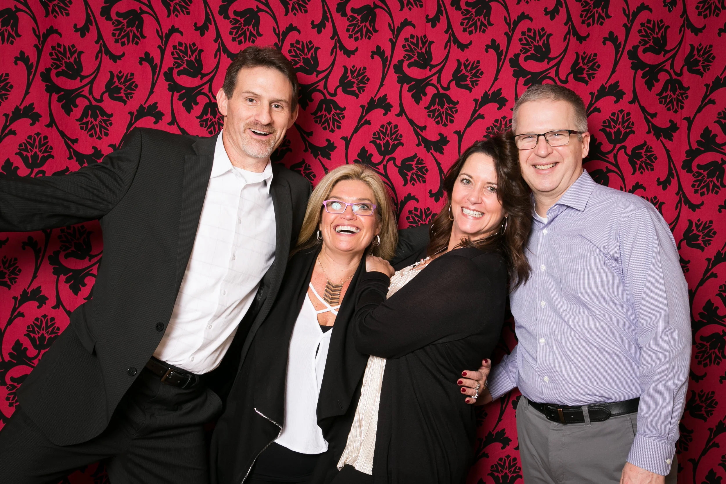 Co-workers in formal business attire posing in front of a red damask backdrop for a corporate party photo lounge rental.