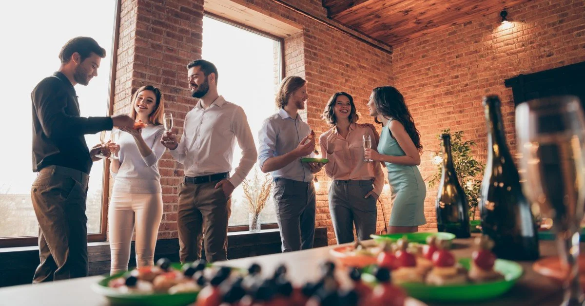 Six people gathered next to an exposed brick wall and windows, with a table of finger foods and wine nearby.