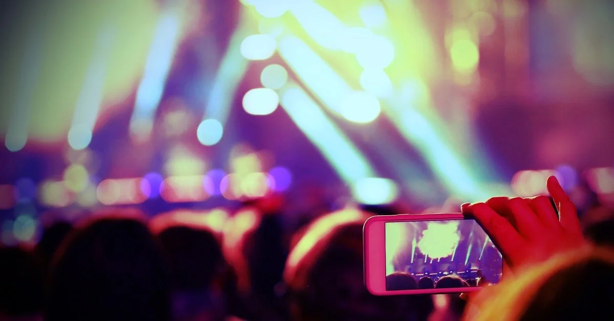 A blurry view of a live event with someone holding their phone up to take a photo for Instagram. The background is dark.