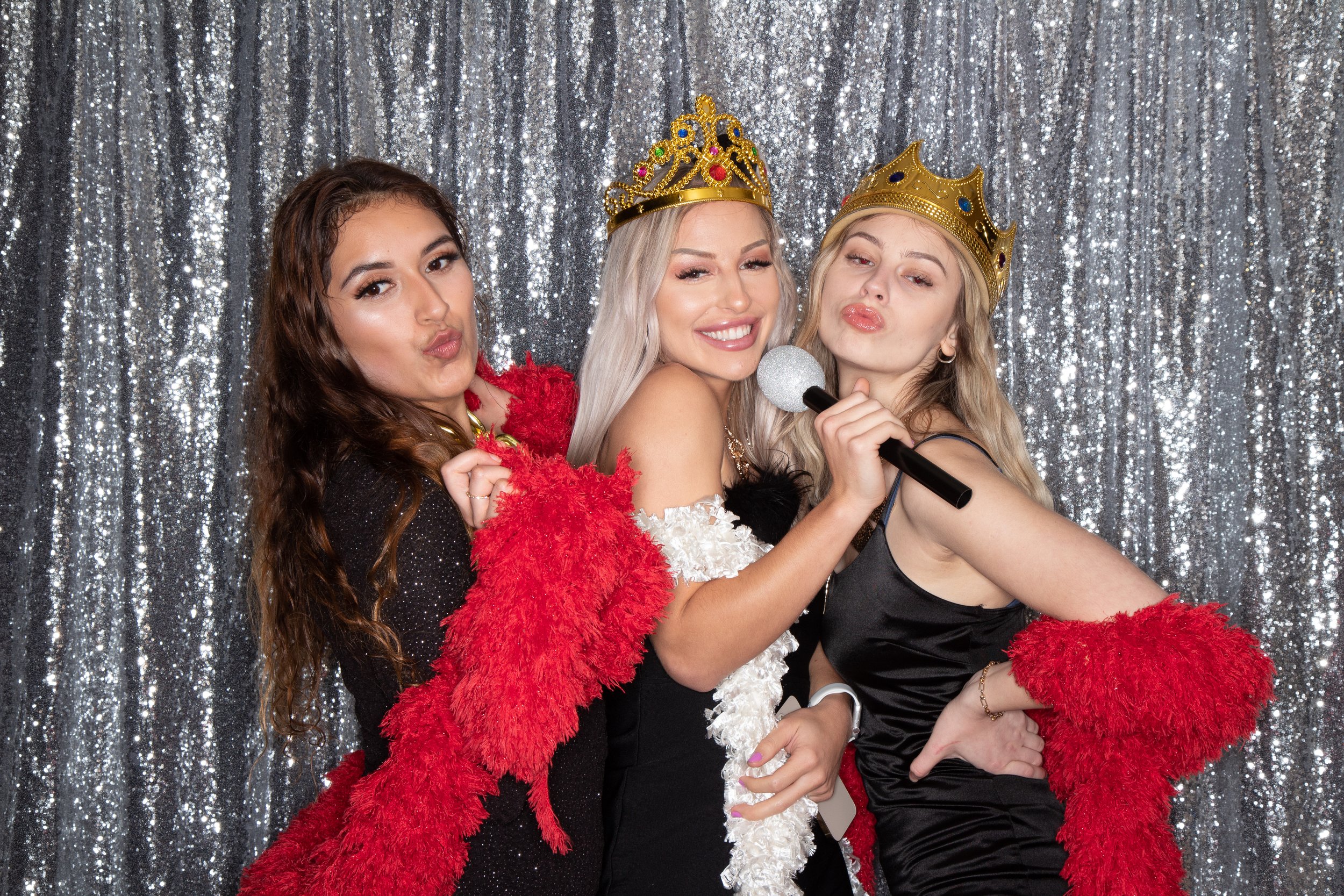 Three women wearing crowns and boa props posing with a microphone against a silver sequin backdrop for the 3-hour Touch Booth Plus package.