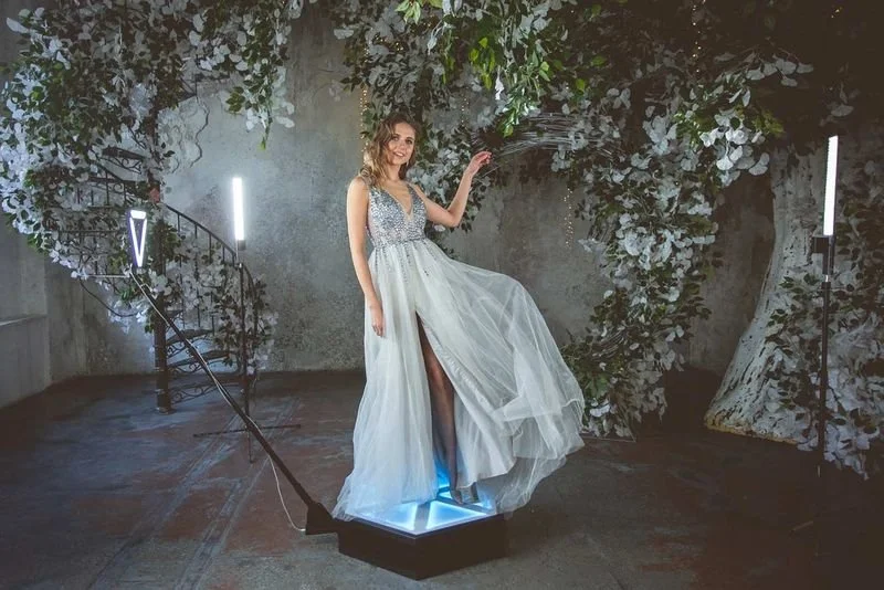 A woman in an elegant blue dress posing on an illuminated 360 video booth platform surrounded by floral decor.