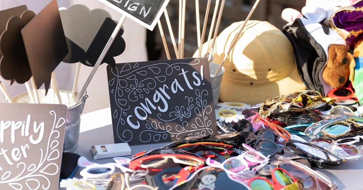 An outdoor event table setup with photo booth props, including signs, glasses, hats, and chalk. One sign says "Congrats!"