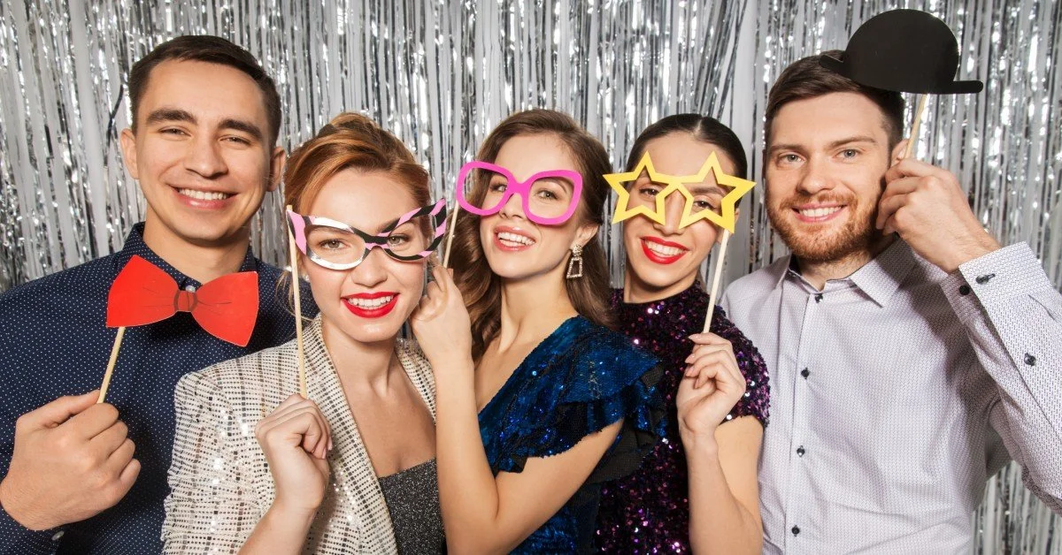 A group of five friends using a photo booth at an event. The backdrop is silver and they're all holding props.