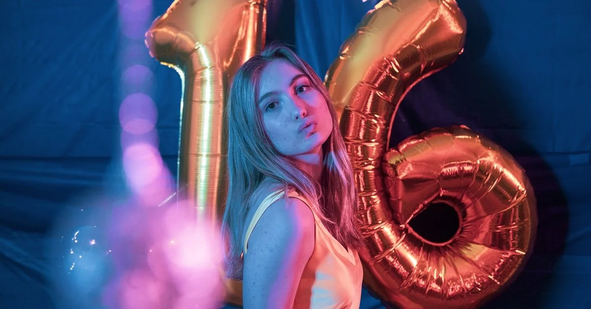 A sweet 16 birthday girl stands in front of balloons in the shape of a 16. She is posing for the photo booth.