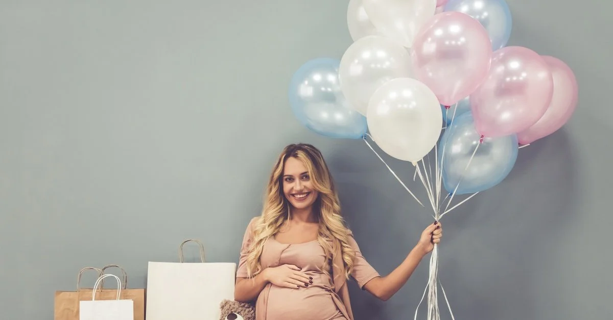 A young pregnant woman in a dress sitting on the floor holding balloons as she poses for baby shower photos.