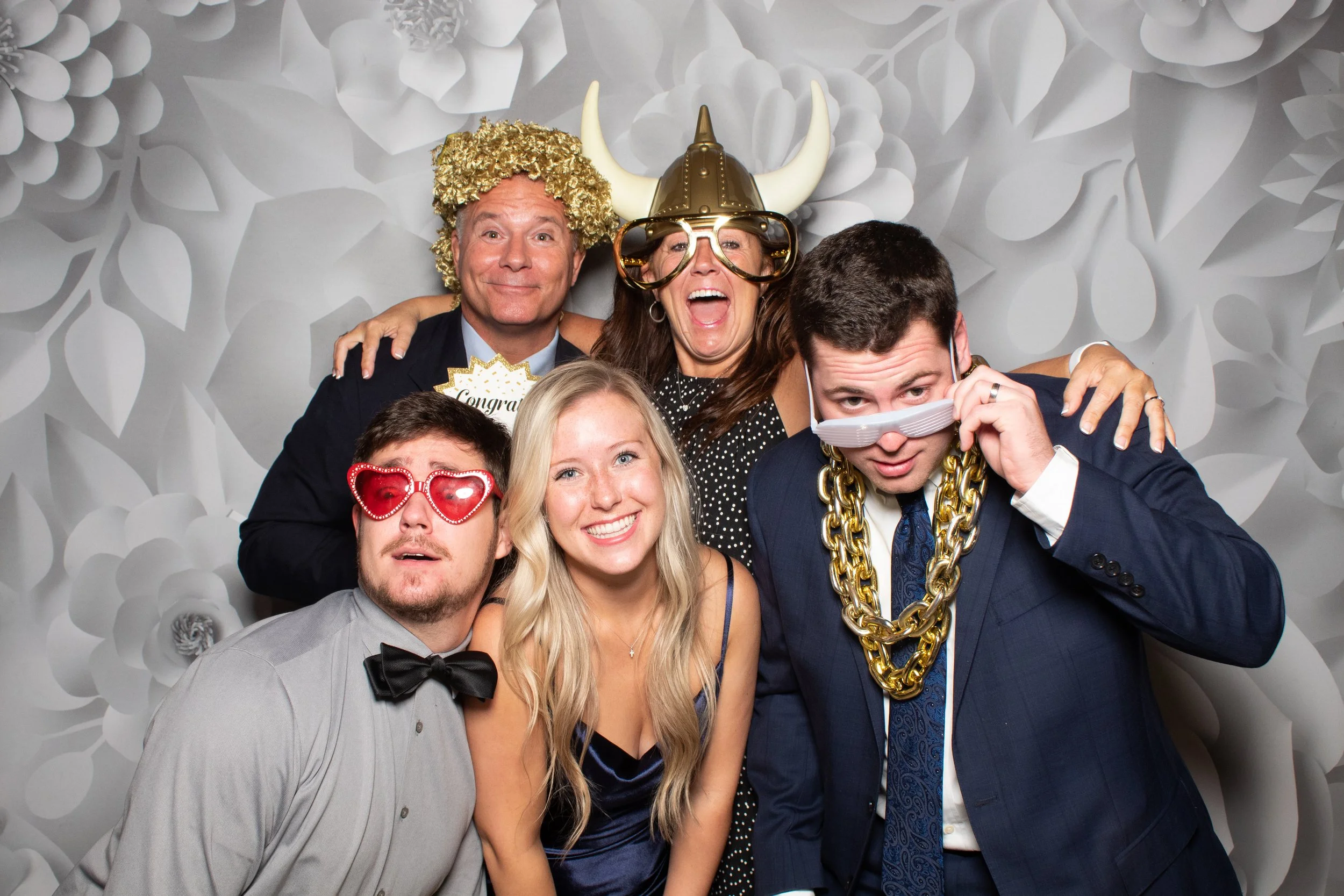 A group of friends wearing fun glasses, hats, and chain props in front of a white 3D floral backdrop for the 4-hour Touch Booth Deluxe package.
