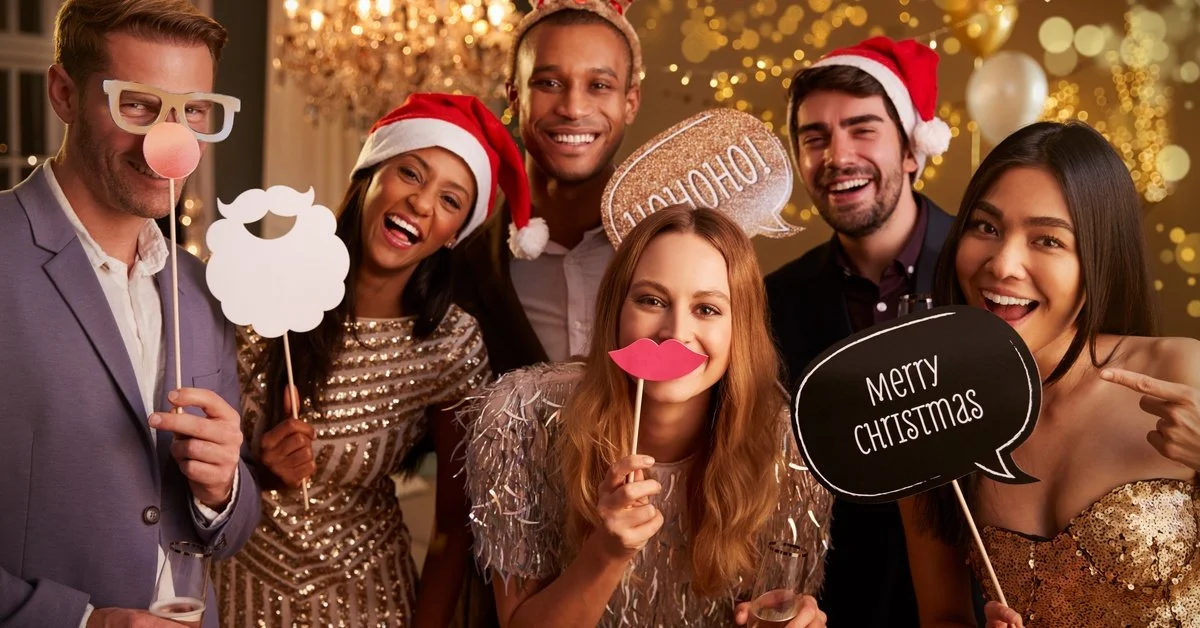 A group of friends at a holiday party stand with props, posed for the photo. One person's sign says "Merry Christmas."