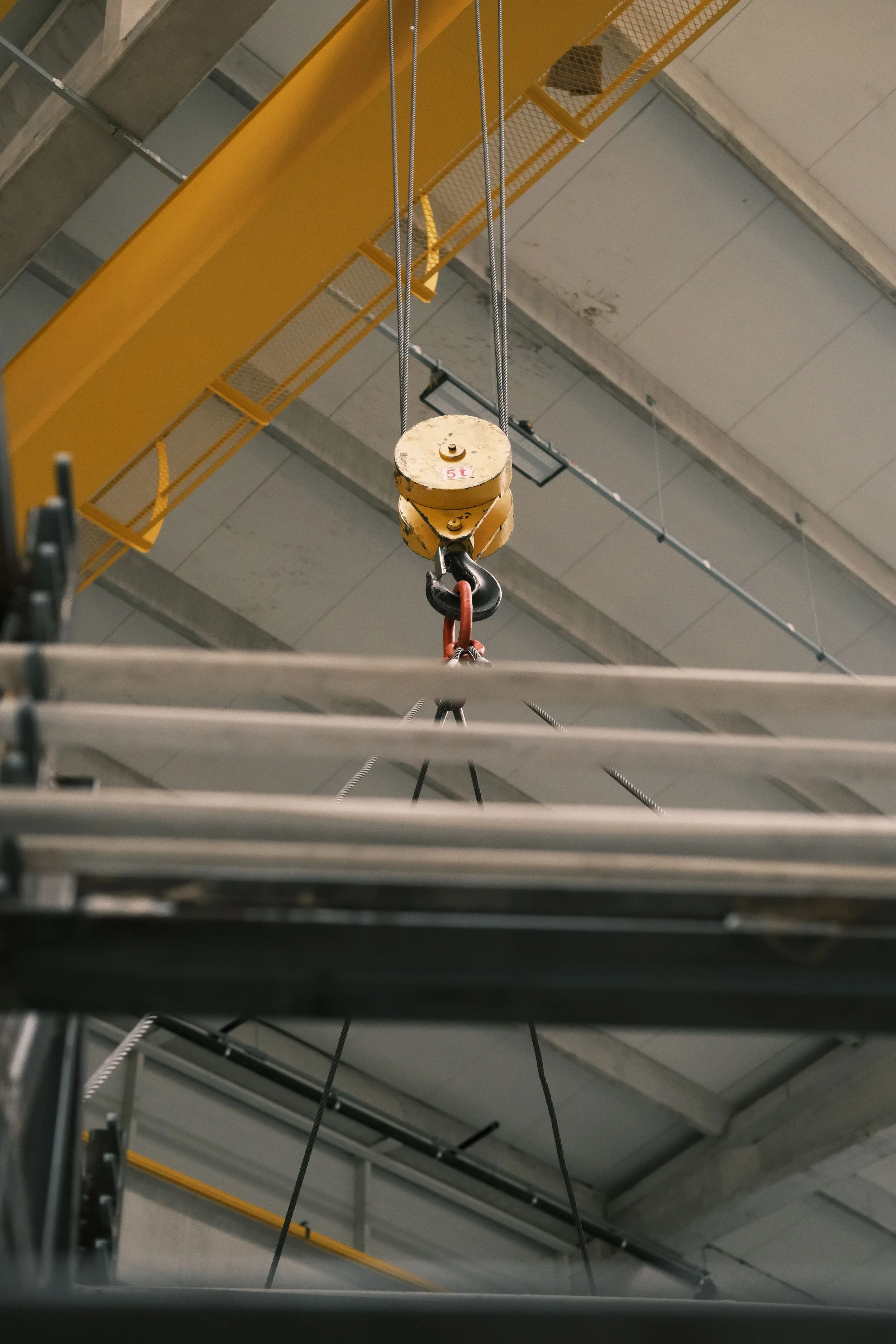 A yellow industrial ceiling hoist with a weight limit label, hanging in a warehouse or industrial setting, with metal beams, pipes, and a yellow safety railing overhead.