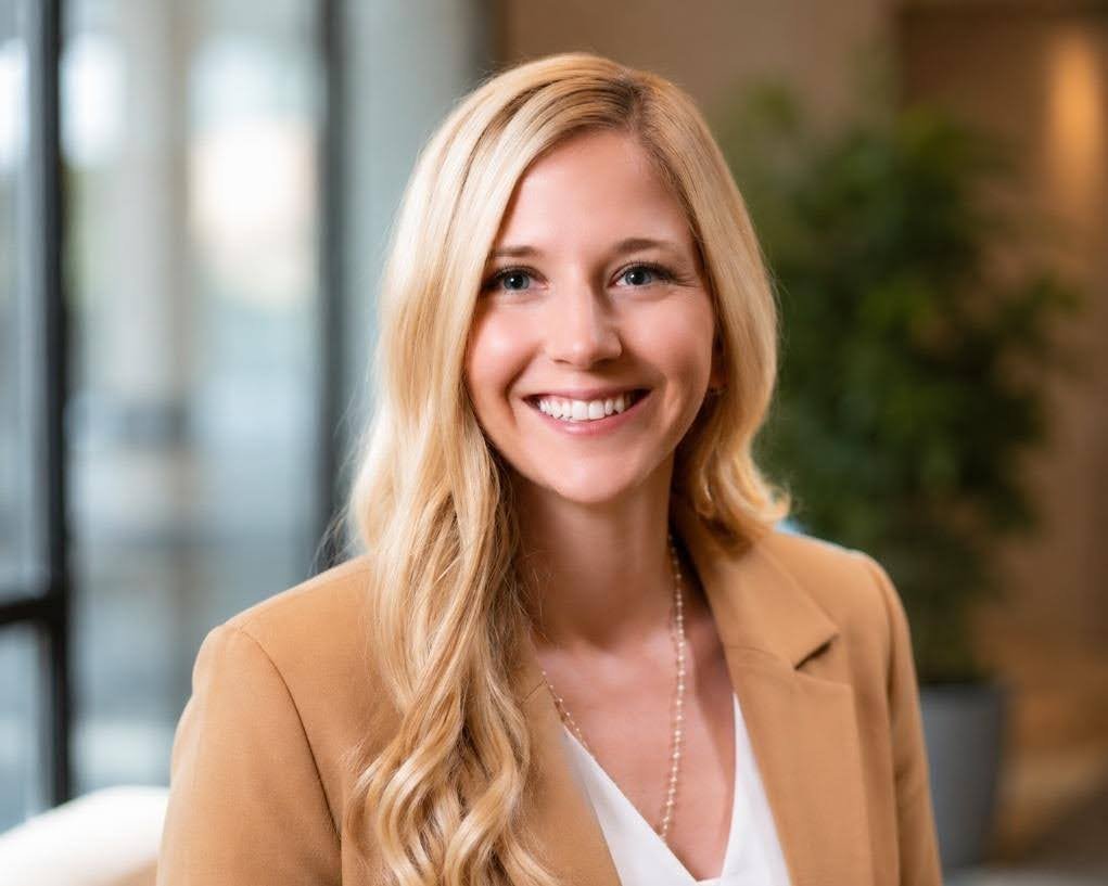 A smiling woman with long blonde hair, wearing a tan blazer, a white top, and a pearl necklace, in an indoor setting with a blurred background of greenery and windows.