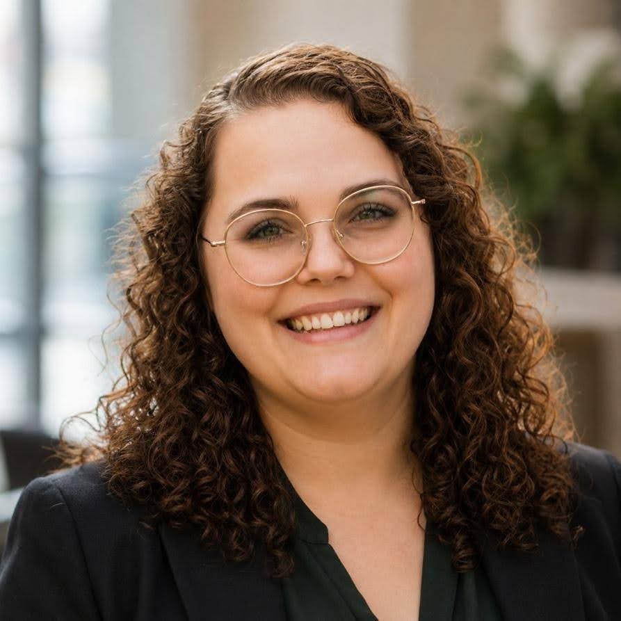 A woman with curly brown hair and glasses smiling in an indoor setting.