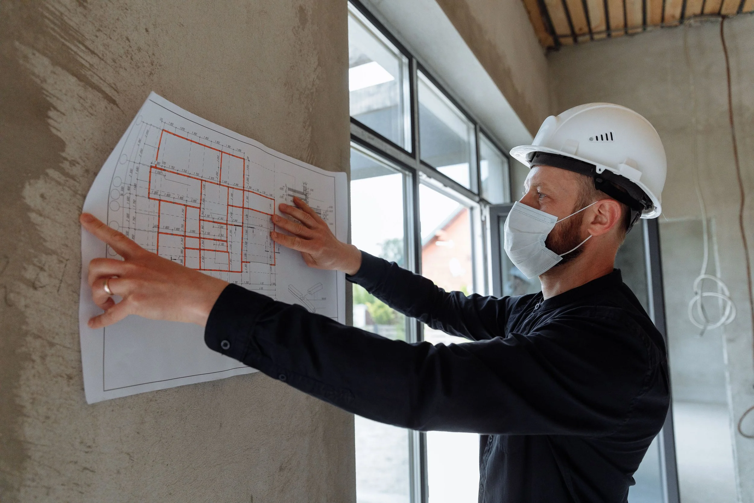 A construction worker wearing a white safety helmet and face mask pointing at a blueprint on a wall inside a building under construction.
