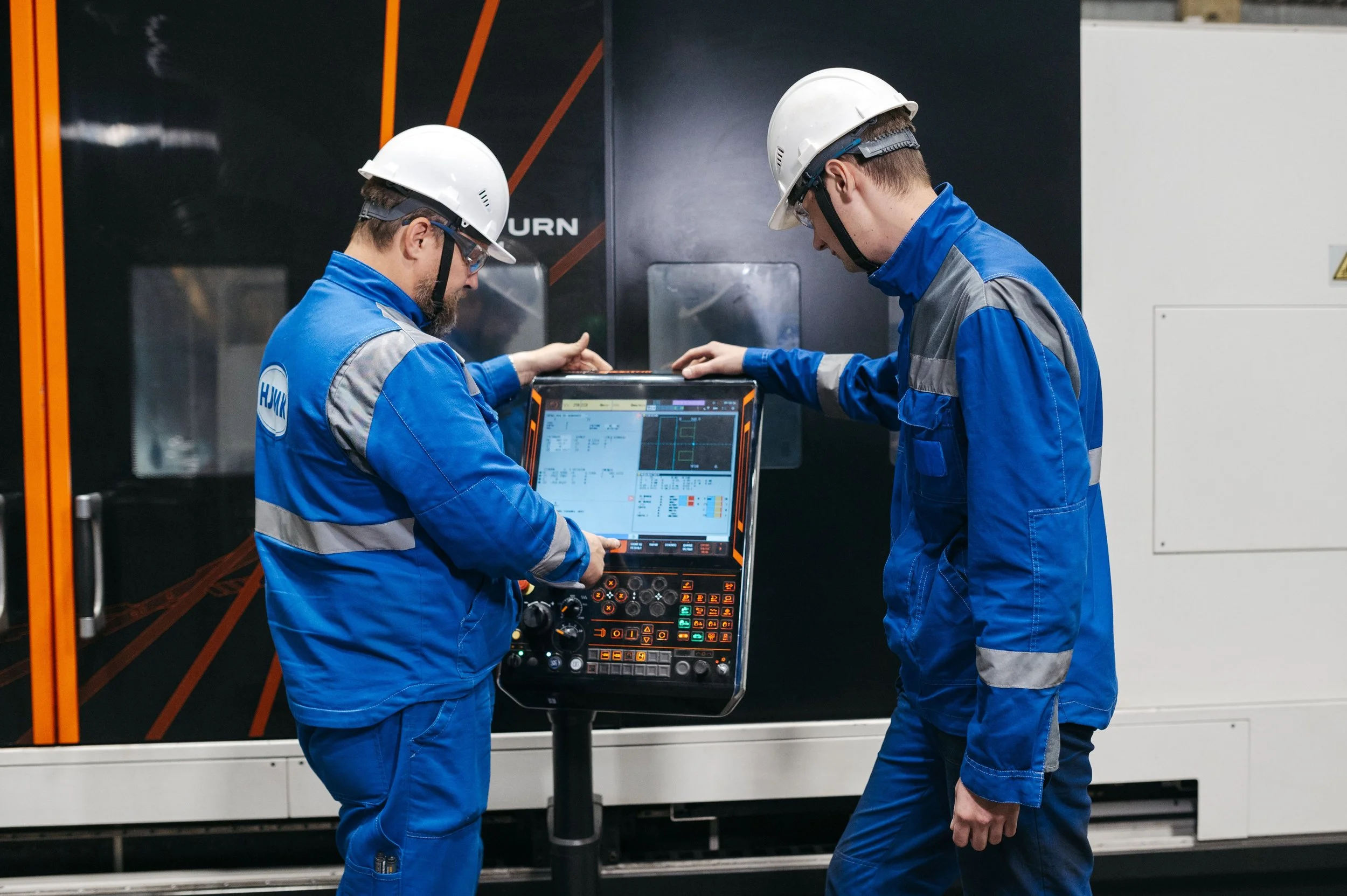 Two male workers wearing blue uniforms, white hard hats, and safety glasses are looking at a digital control panel in an industrial setting, possibly a manufacturing or power plant facility.