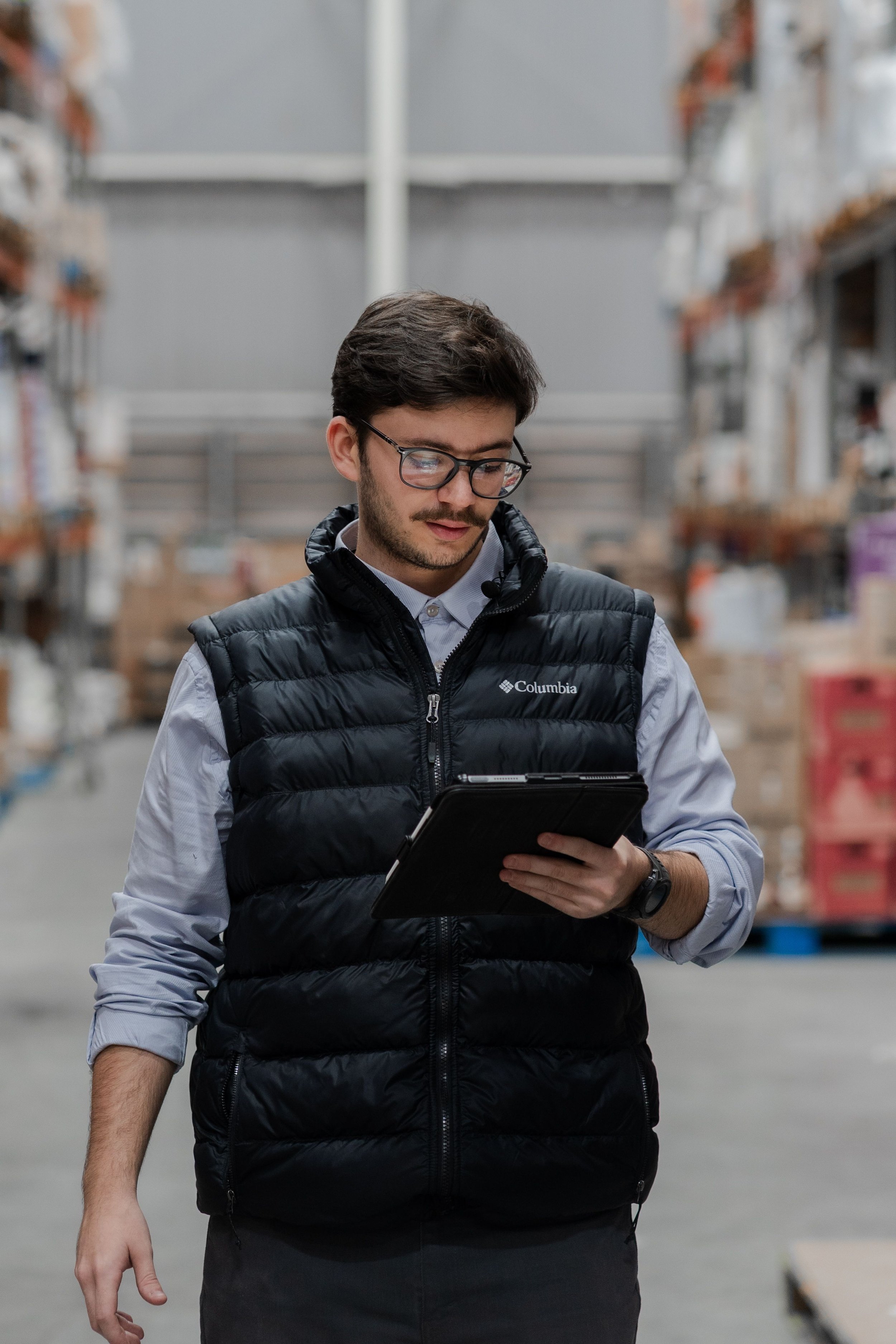 A man wearing glasses and a black Columbia puffer vest over a long-sleeve shirt, looking at a tablet, in a warehouse with shelves of boxes.