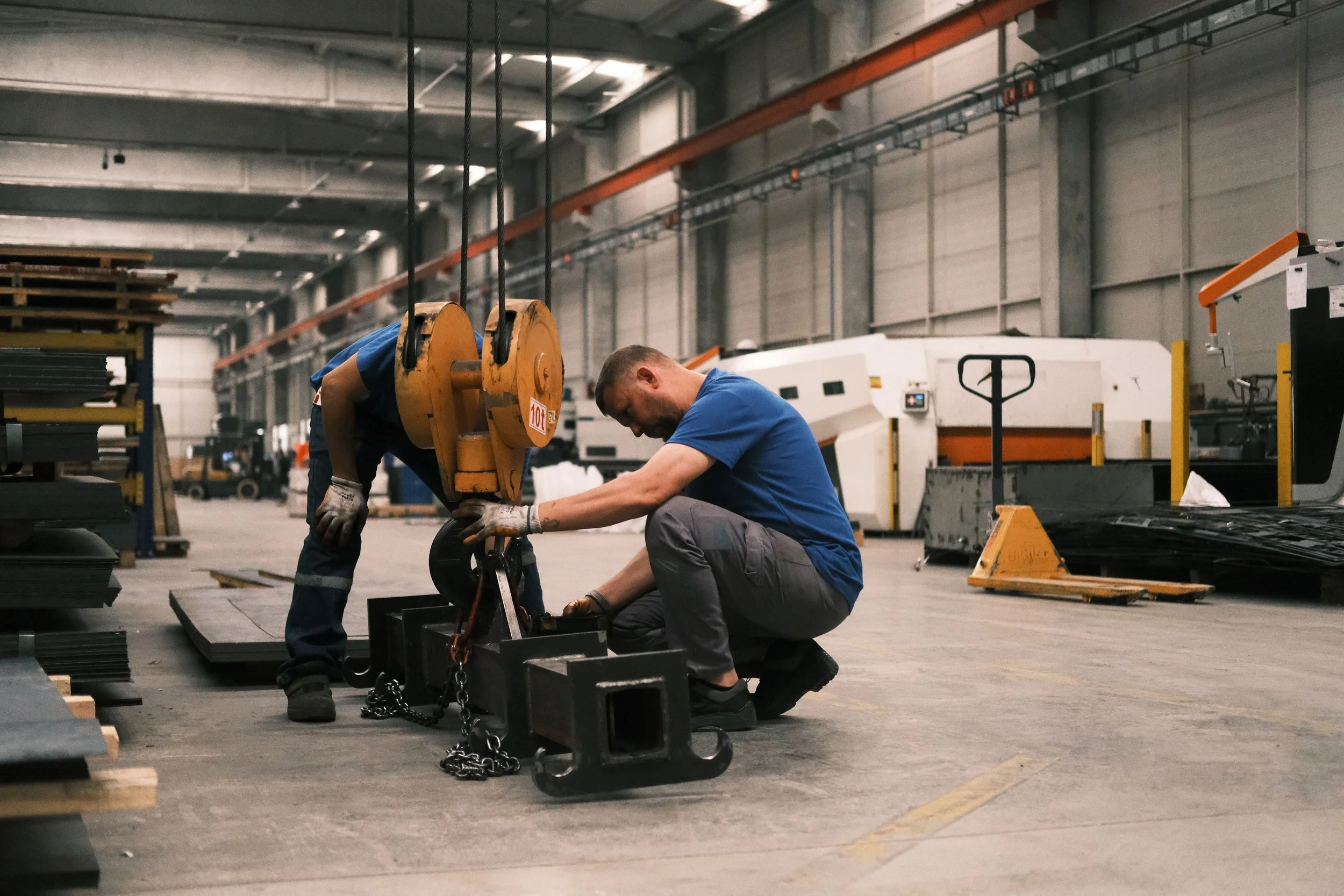Two workers in a factory or warehouse are working on a metal structure, with one kneeling and the other bent over, using tools. They are surrounded by industrial equipment and materials, with shelves and machinery visible in the background.
