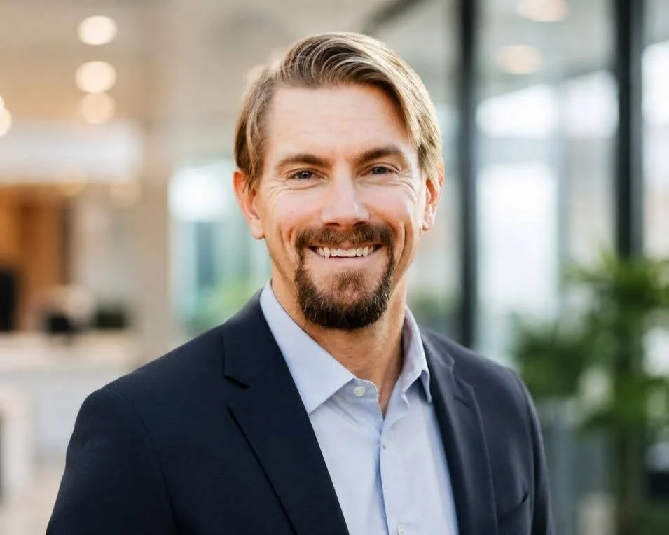 A smiling man with blonde hair and a beard wearing a dark suit jacket and light blue collared shirt, standing in a modern office with large windows and blurred interior background.