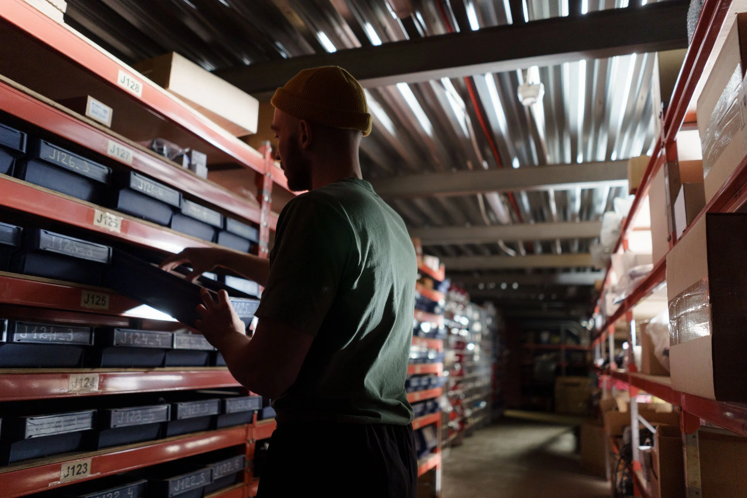 A man wearing a yellow beanie and a green t-shirt shopping in a warehouse with shelves filled with labeled boxes.