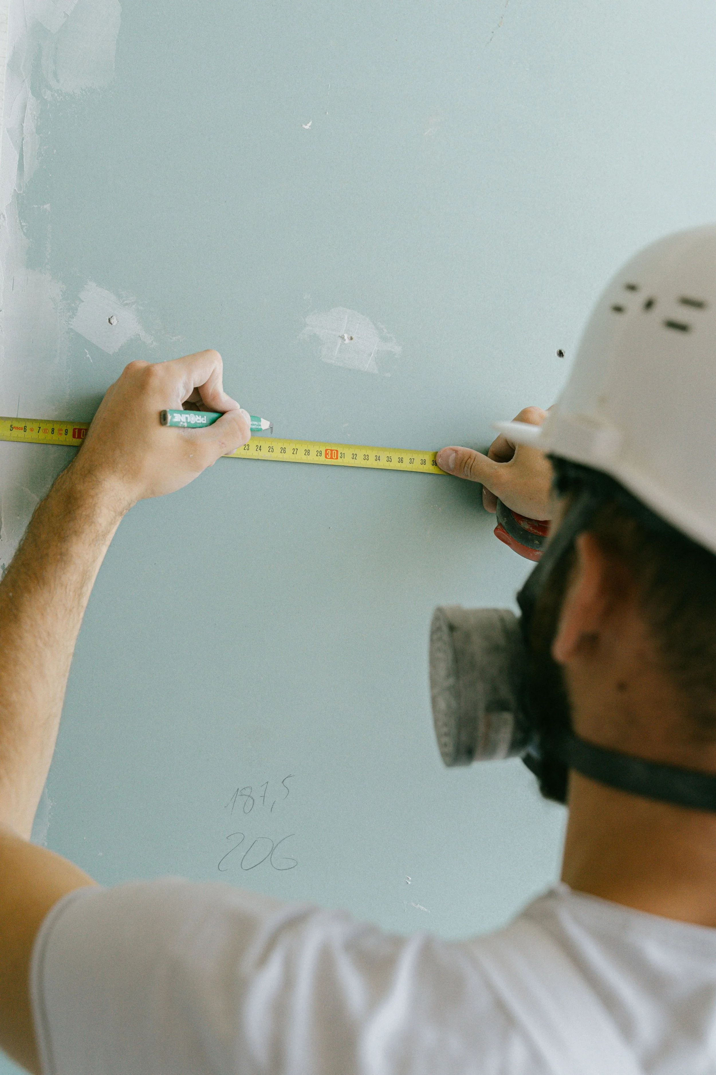 A person wearing a white hard hat is measuring a light blue wall with a tape measure, while another person's hand writes measurements and notes on the wall.