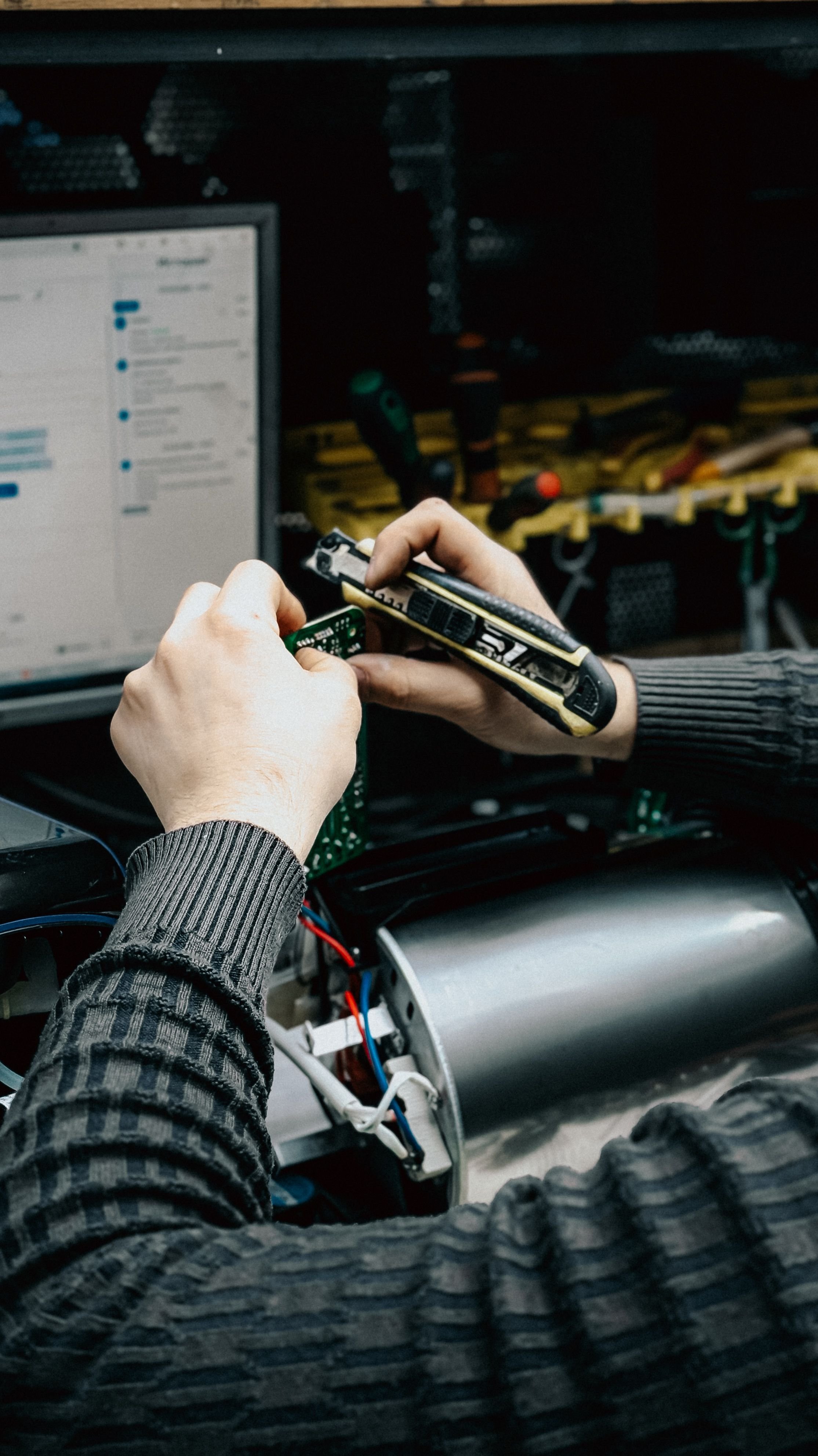 Person working on electronic circuit in a workshop, with tools and a computer monitor in the background.