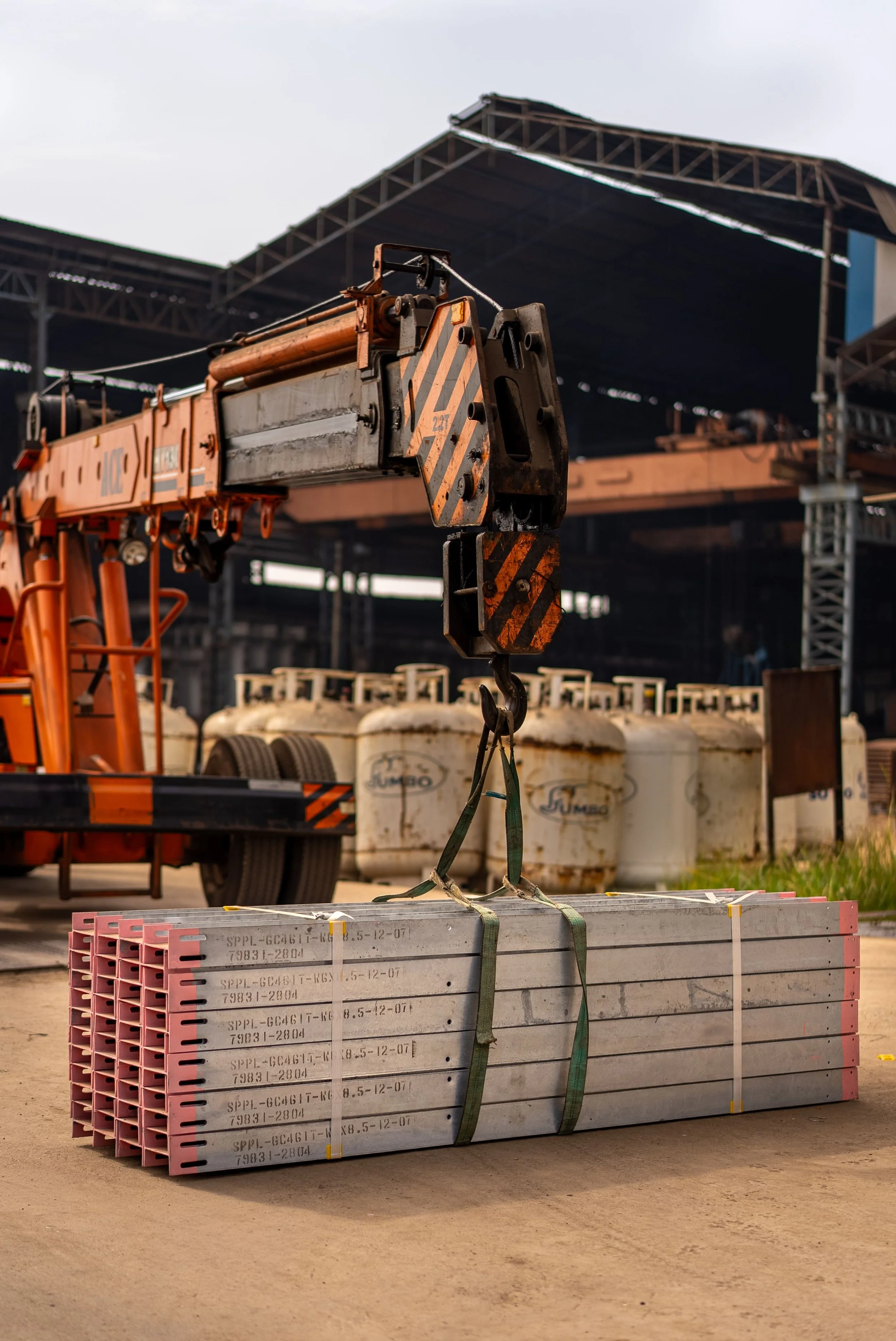 A construction site with a crane lifting a large metal panel or framework, with other construction equipment and containers in the background.