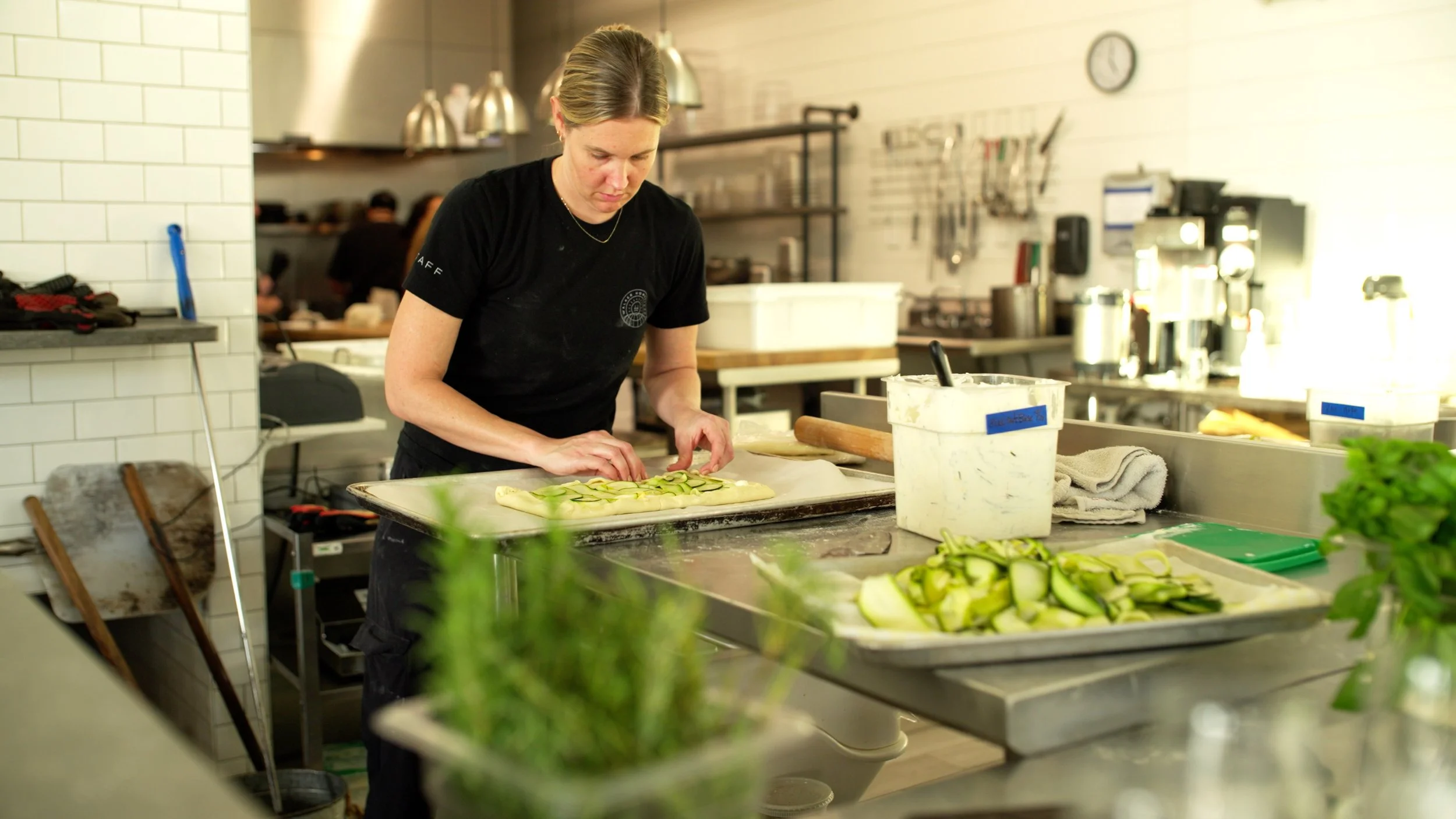 A woman preparing sliced vegetables on a tray in a professional kitchen with various kitchen tools and appliances in the background.