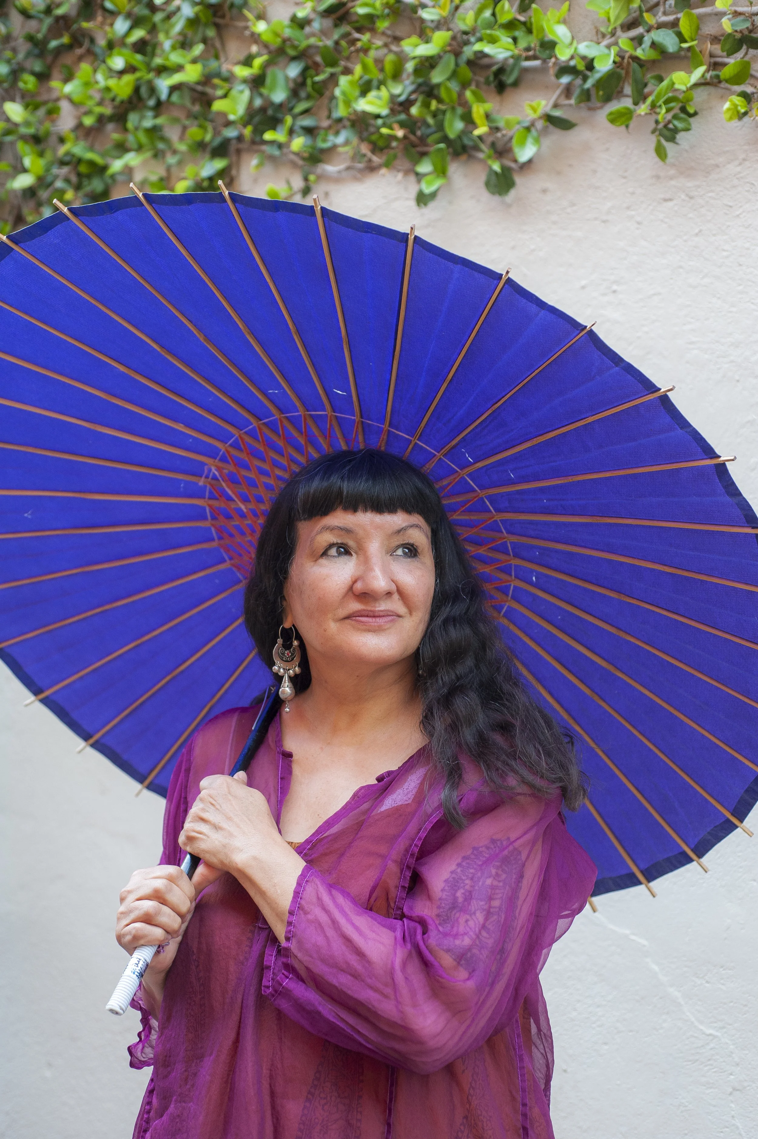 A dark haired woman is wearing a purple top and carrying a blue umbrella. She stands in front of a white wall with green foliage hanging above her.
