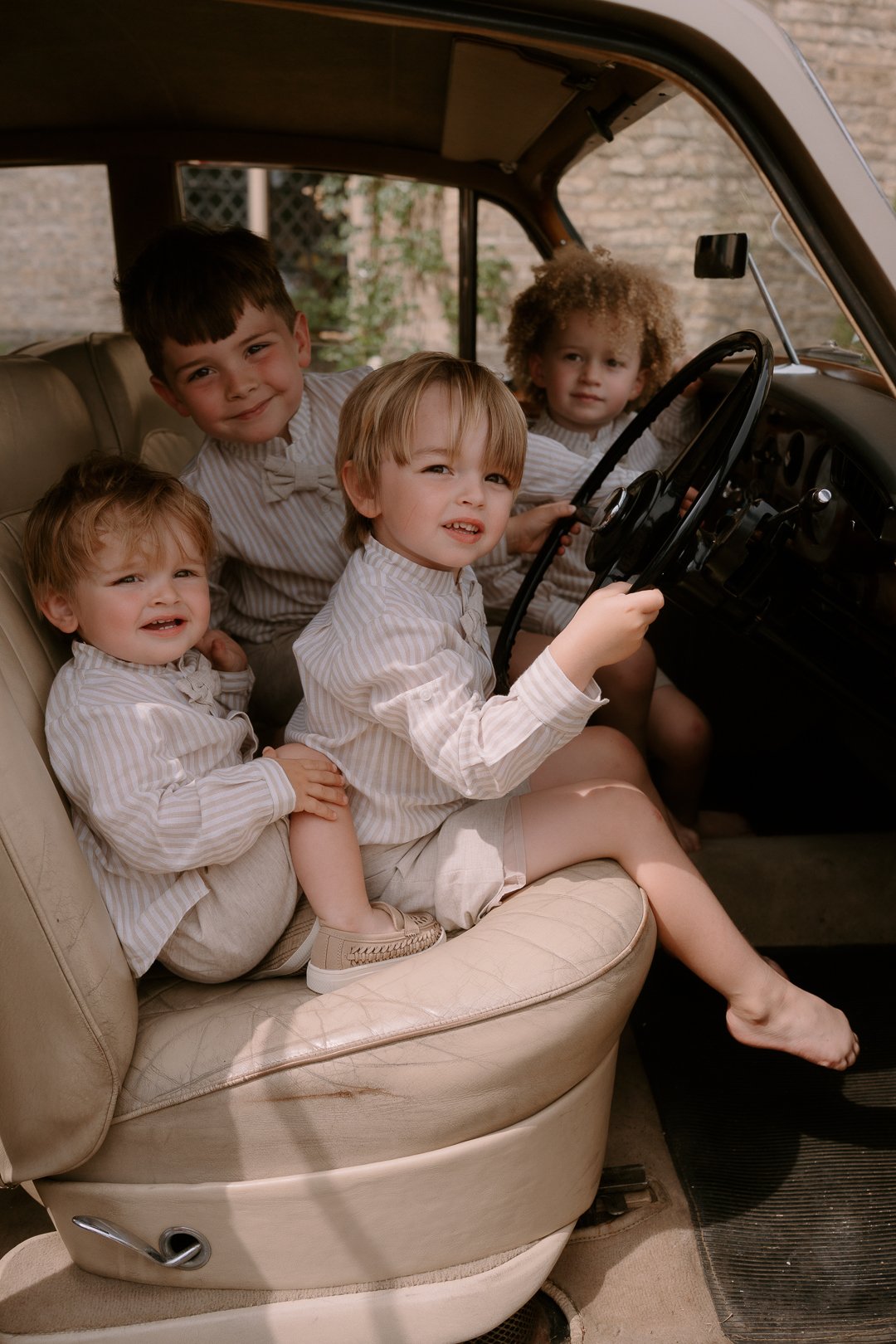 Five young children sitting inside a vintage car, wearing light-colored striped shirts, with one child holding the steering wheel.