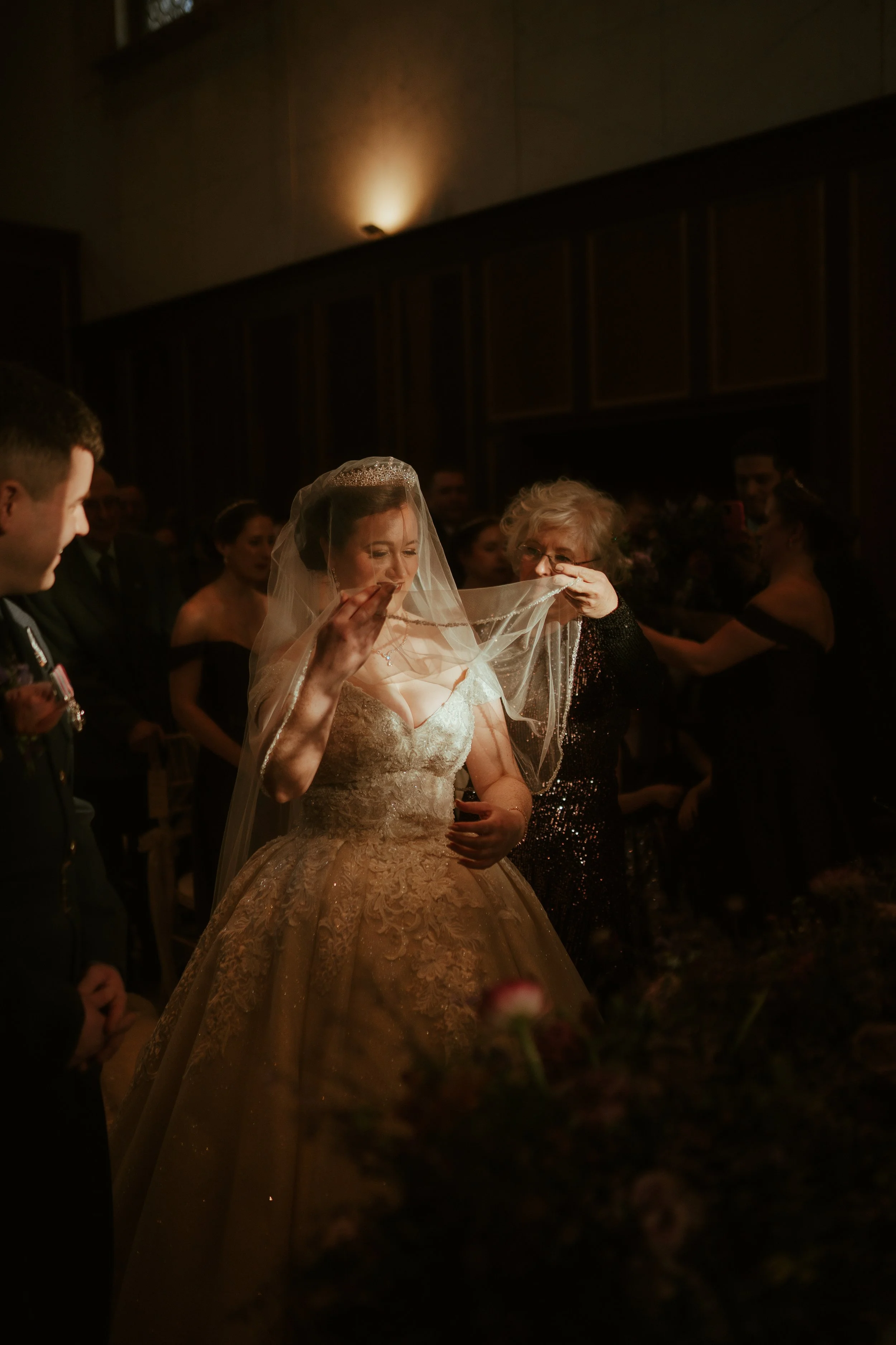 A bride in a wedding gown with lace details and a veil, receiving a toast or blessing from an elderly woman at a wedding reception. Several guests are visible in the background.