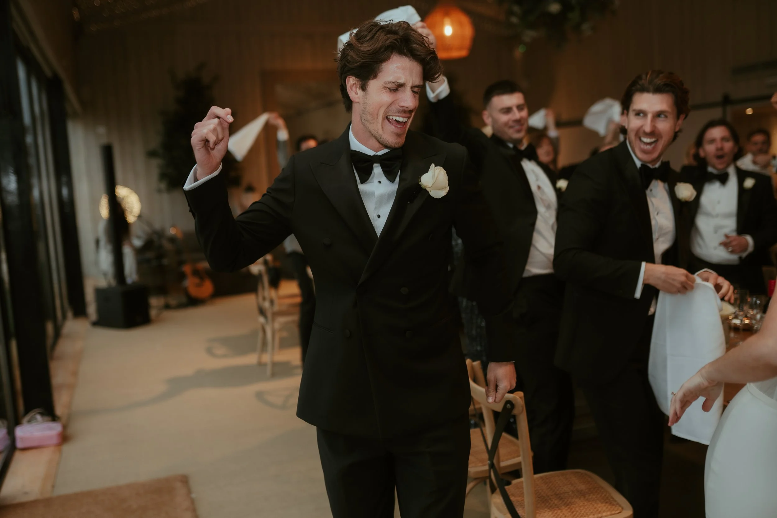 A groom in a black tuxedo and bow tie is dancing and smiling with his eyes closed at a wedding reception, surrounded by other guests also dressed in formal attire, in a warmly lit indoor venue.