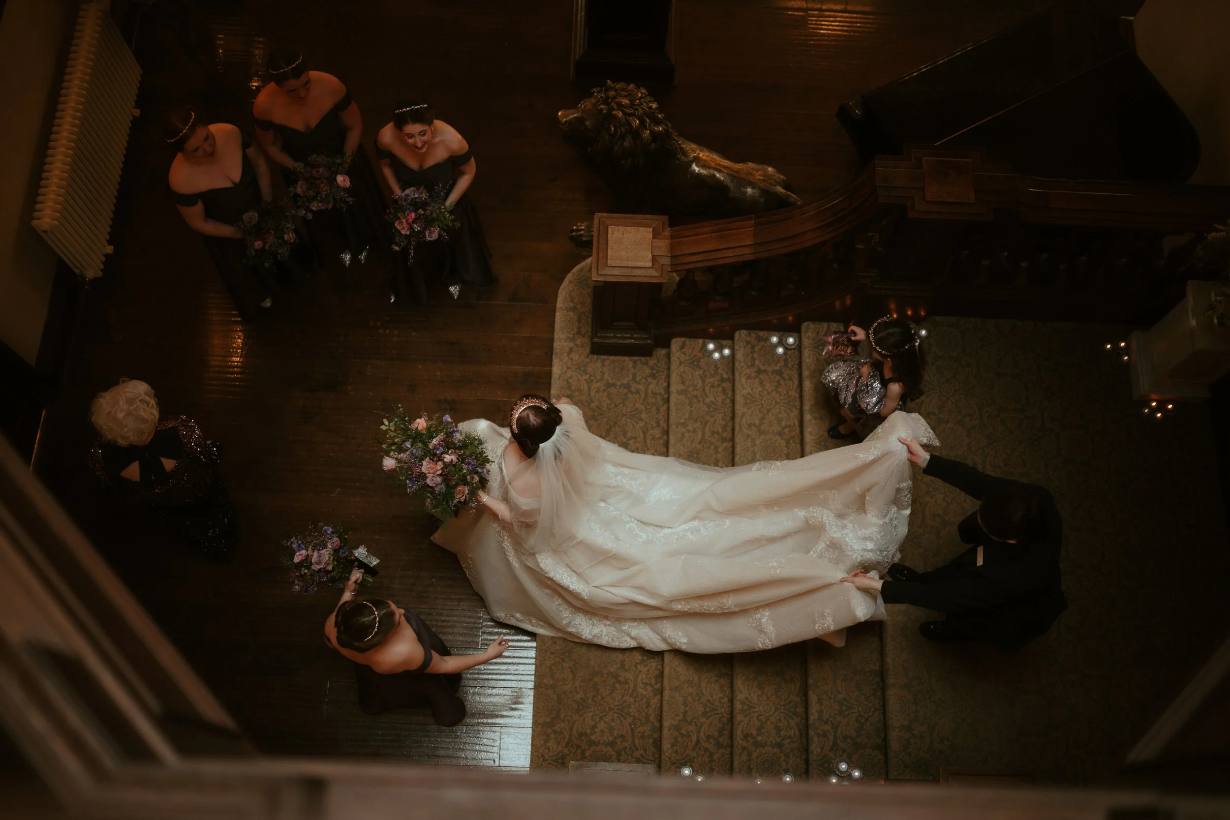 Top-down view of a bride in a white gown with a long train, holding a bouquet, surrounded by attendants in black dresses, and some holding bouquets, bourton hall