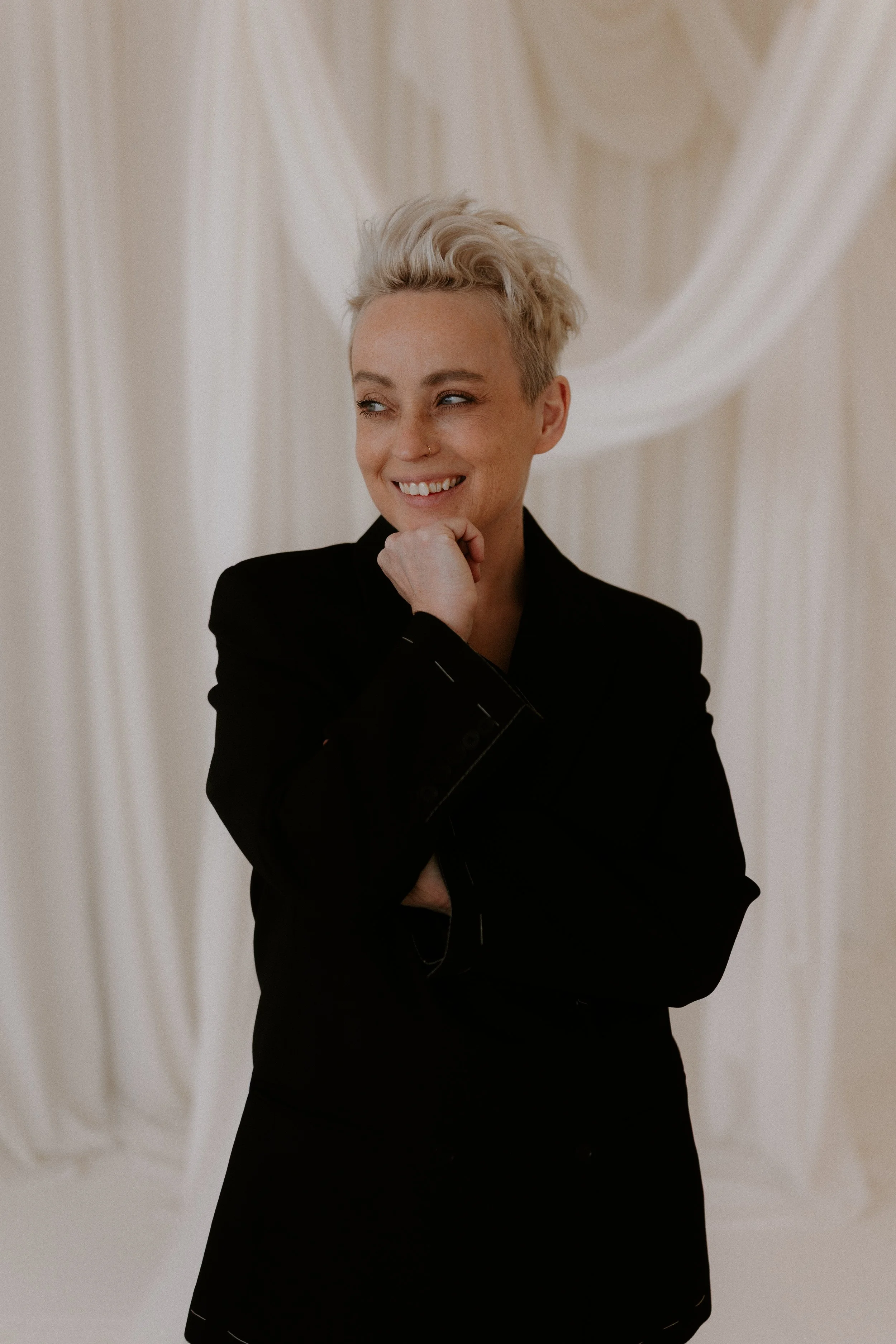 A woman with short blonde hair, smiling and looking to the side, wearing a black jacket, standing in front of a cream-colored curtain backdrop.