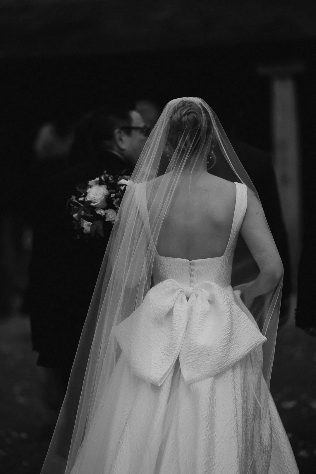 A bride in a wedding dress and veil facing away, holding a bouquet, during her wedding ceremony.