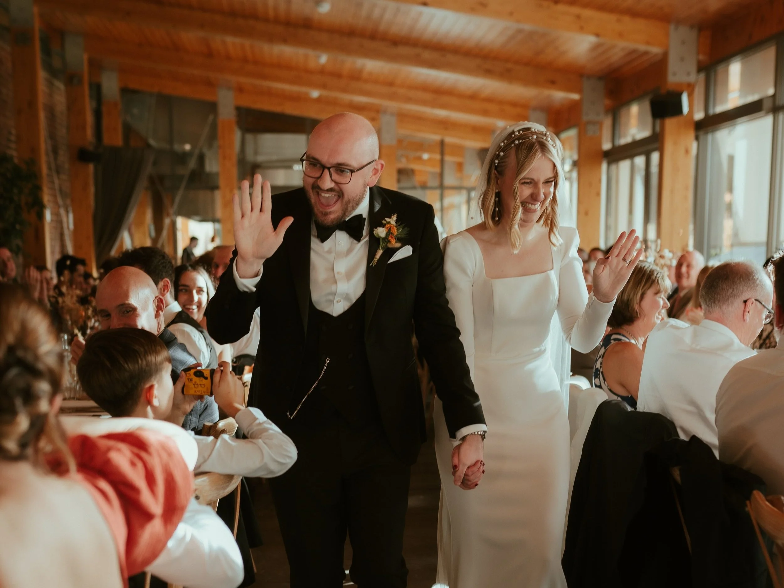 A newlywed couple holding hands and waving at their wedding reception. The groom is wearing a black tuxedo with a bow tie, and the bride is in a white gown with a pearl headband. They are smiling and walking through a crowded venue with wooden beams 