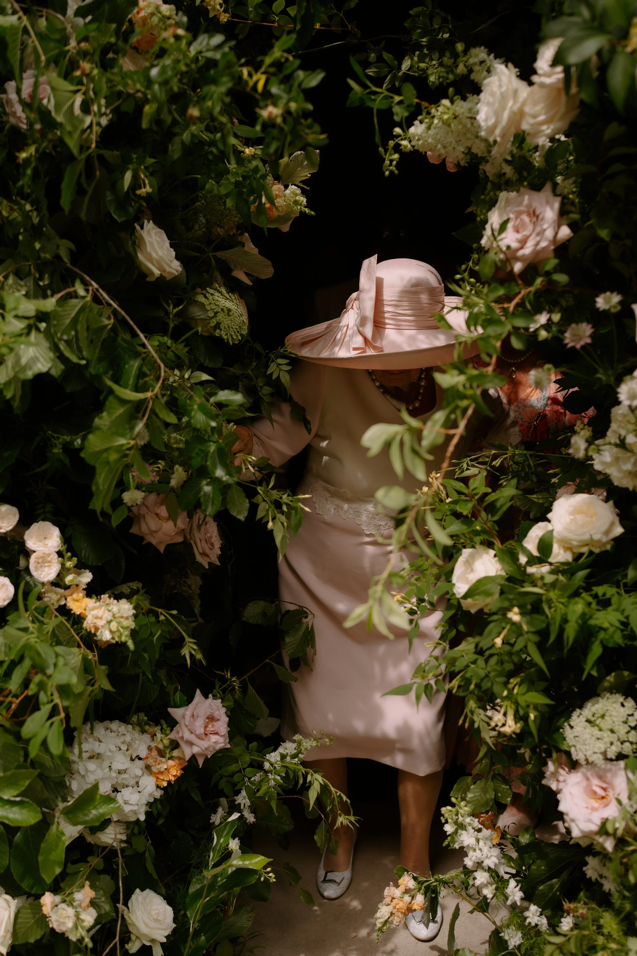 Person wearing a pink wide-brimmed hat and a beige dress, standing among lush green foliage and white flowers, with only part of their face visible.