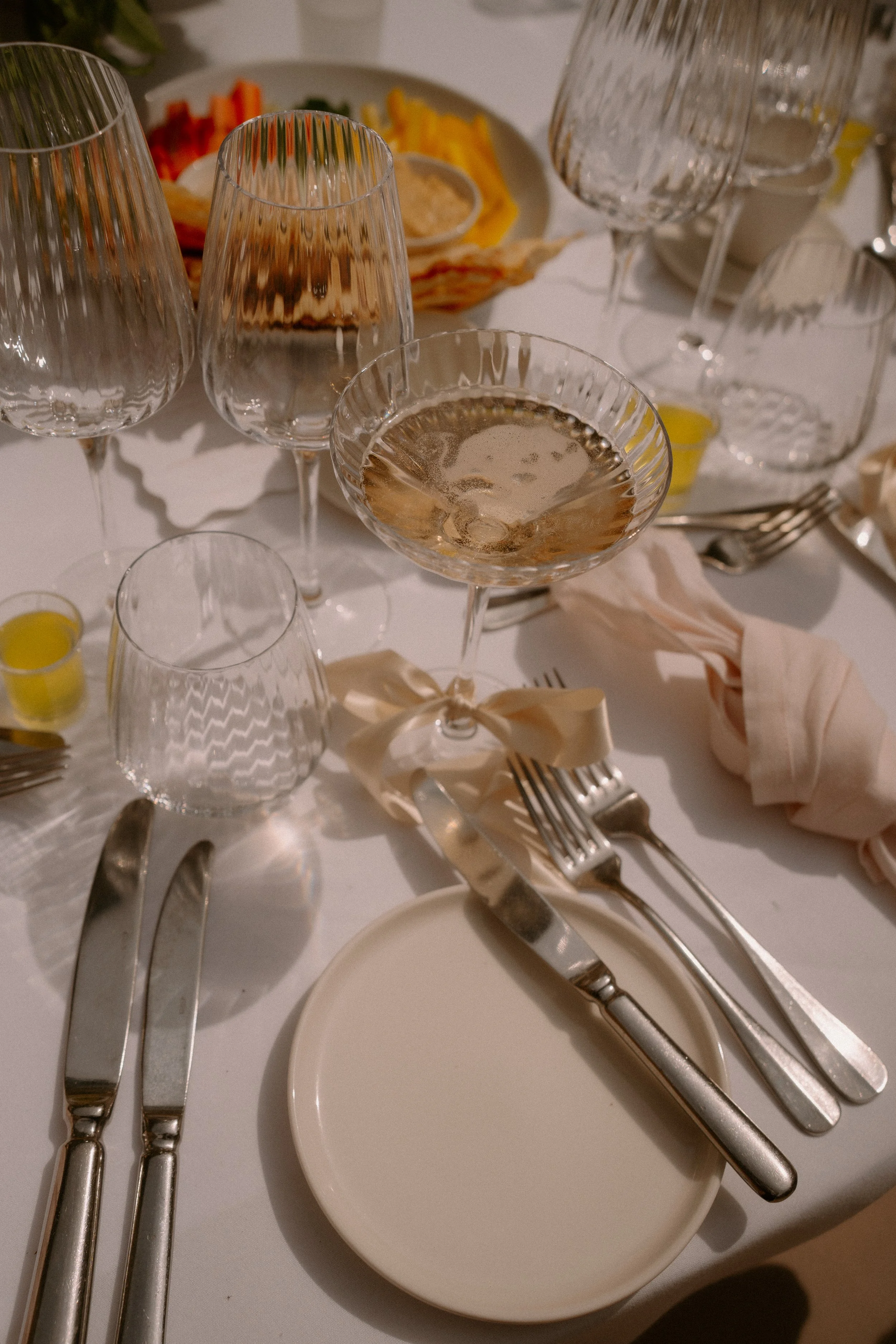 Empty glassware, silverware, a small white plate, and a communal dinner plate with food remnants on a white tablecloth, suggesting a recently finished meal at a formal or semi-formal gathering.