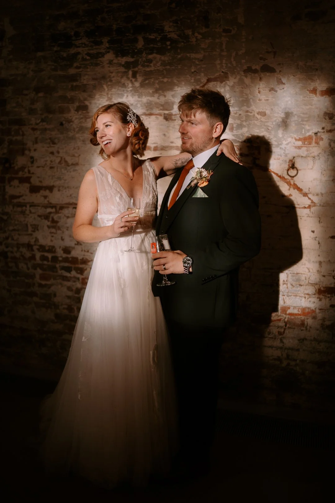 A bride and groom are smiling and enjoying a celebration, standing close together with the bride’s arm around the groom's shoulder. The bride is wearing a white wedding dress and holding a glass of champagne, and the groom is dressed in a black suit 