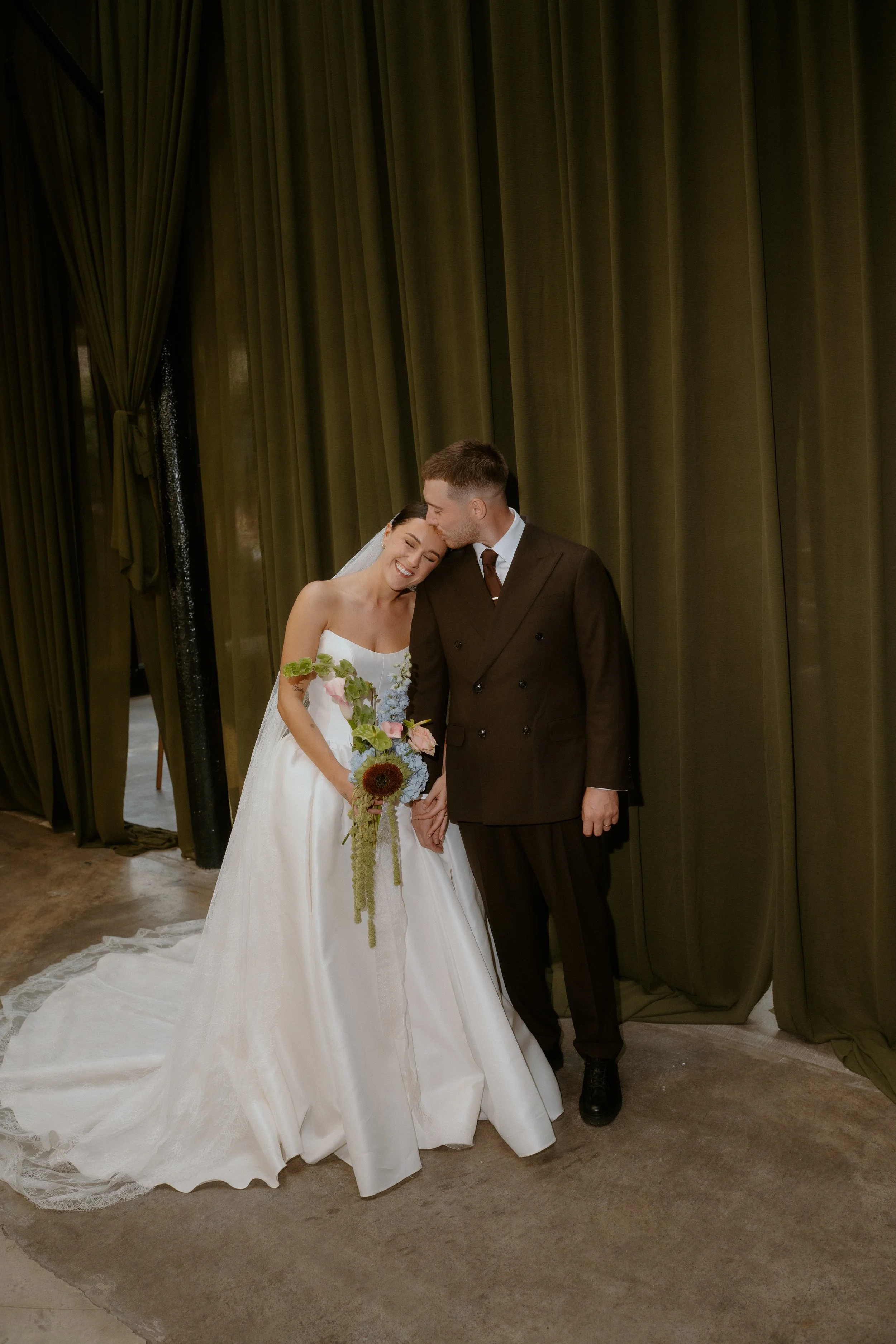 A bride and groom standing close together, the bride is holding a bouquet of flowers, she is smiling, and the groom is kissing her forehead. They are indoors with green curtains in the background.