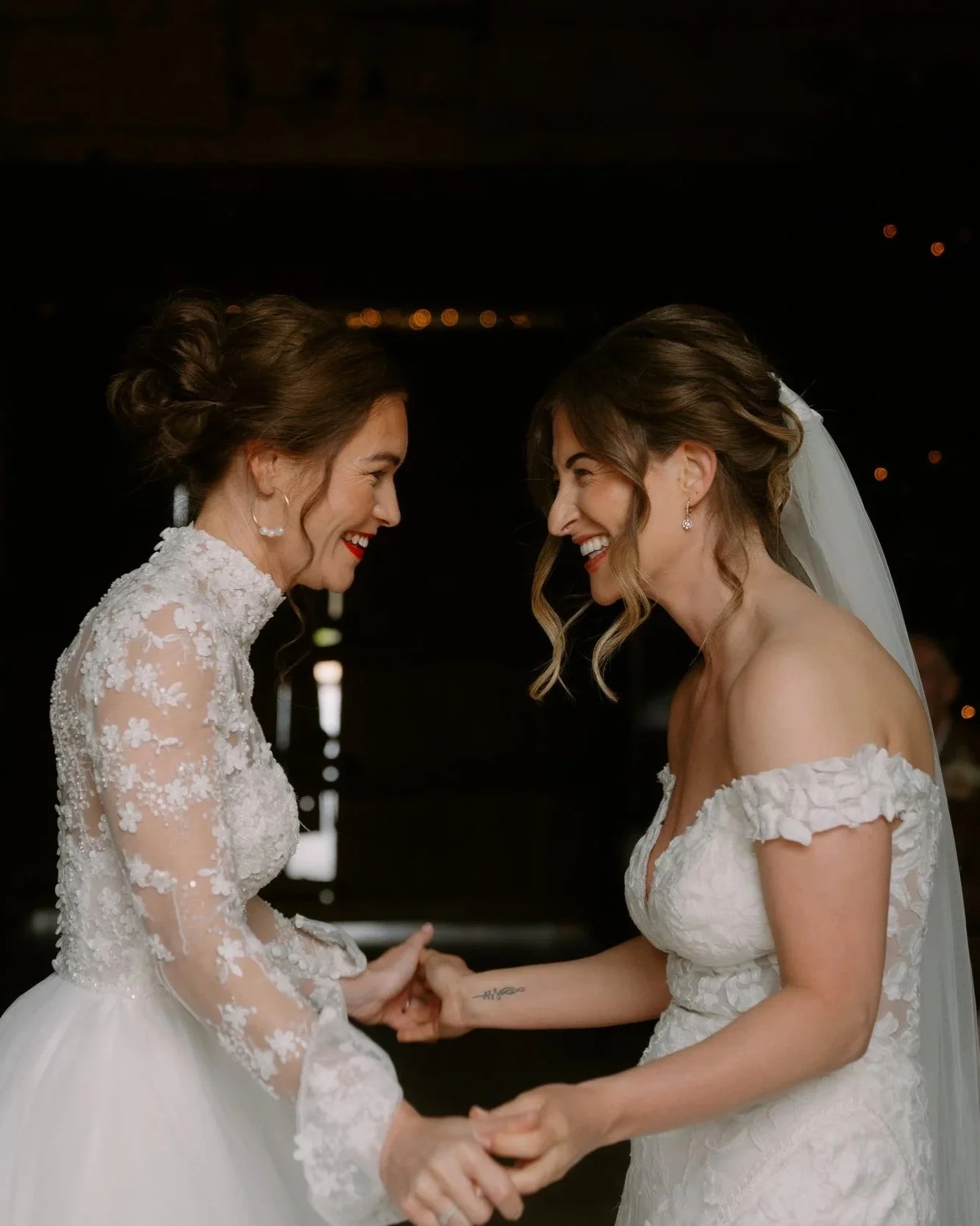 Two women dressed in wedding gowns holding hands and smiling at each other during a wedding ceremony.