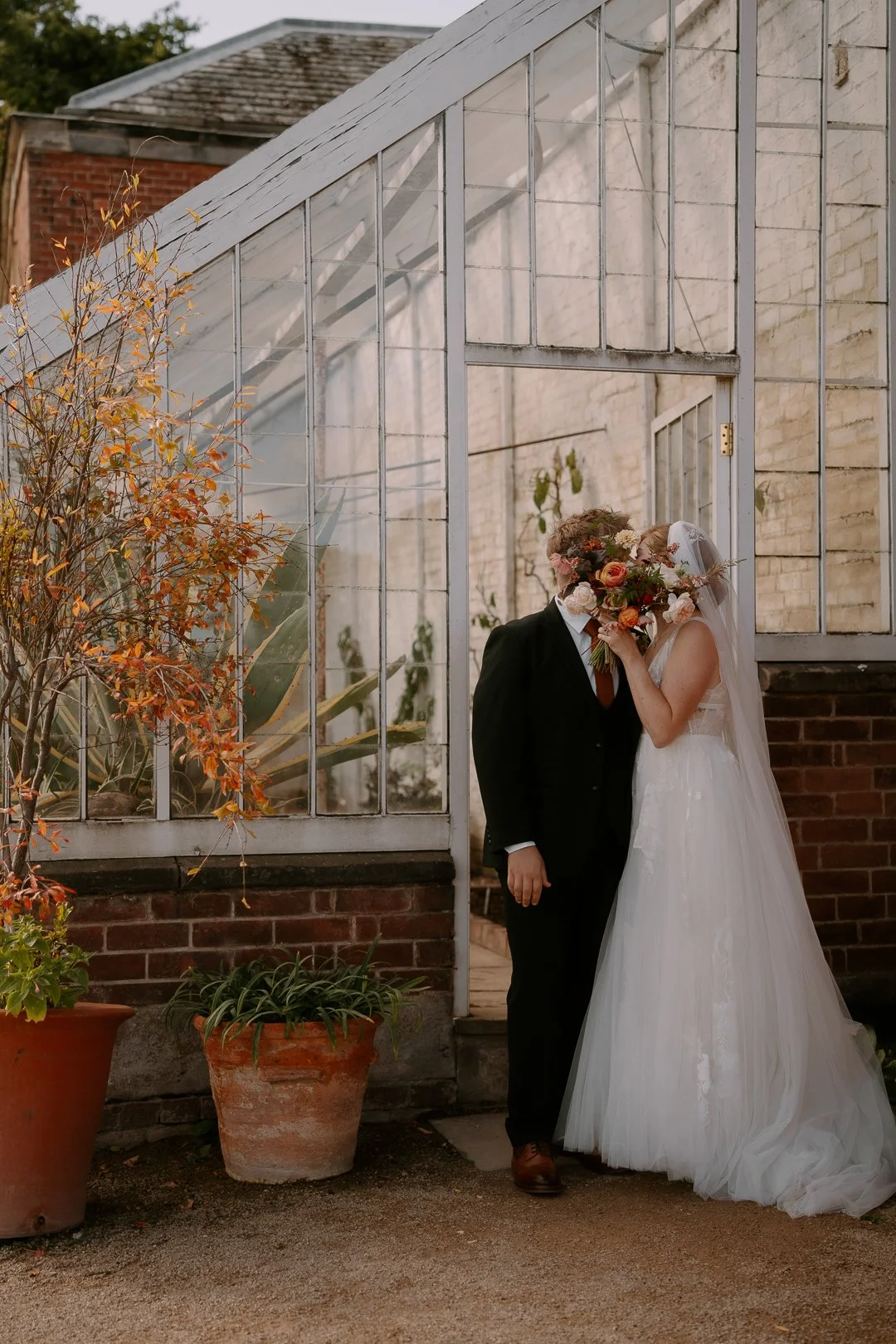 A bride and groom kissing inside a greenhouse, with a large bouquet covering the bride and groom's faces. The bride wears a white wedding dress and veil, while the groom is dressed in a black suit with a white shirt and brown shoes. There are potted 