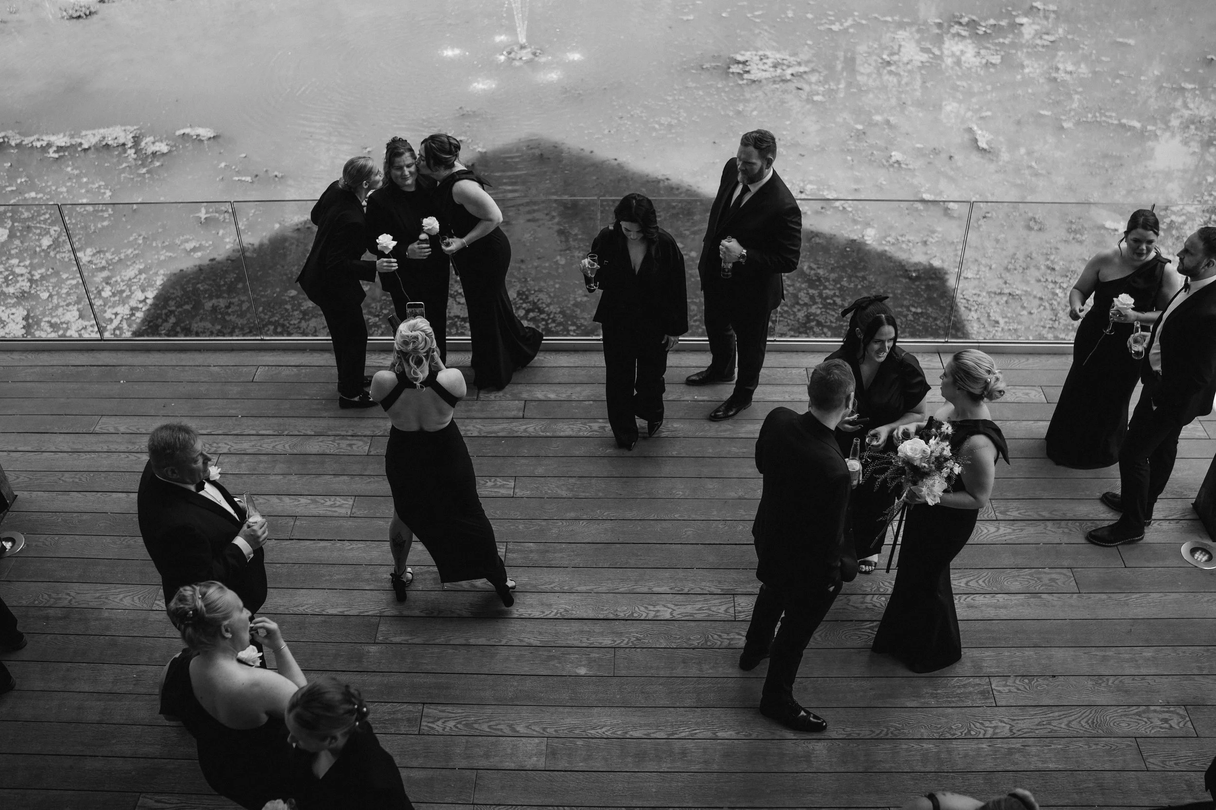 Black and white photo of a wedding reception with guests in formal attire standing on a wooden floor, talking and holding drinks, with a glass barrier and water in the background.