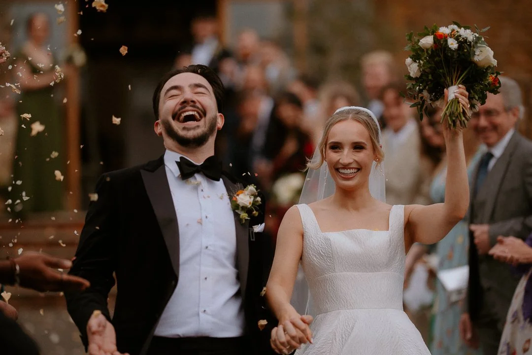 A newlywed couple celebrating their wedding, with the bride holding a bouquet and the groom laughing, surrounded by guests throwing confetti outdoors.
