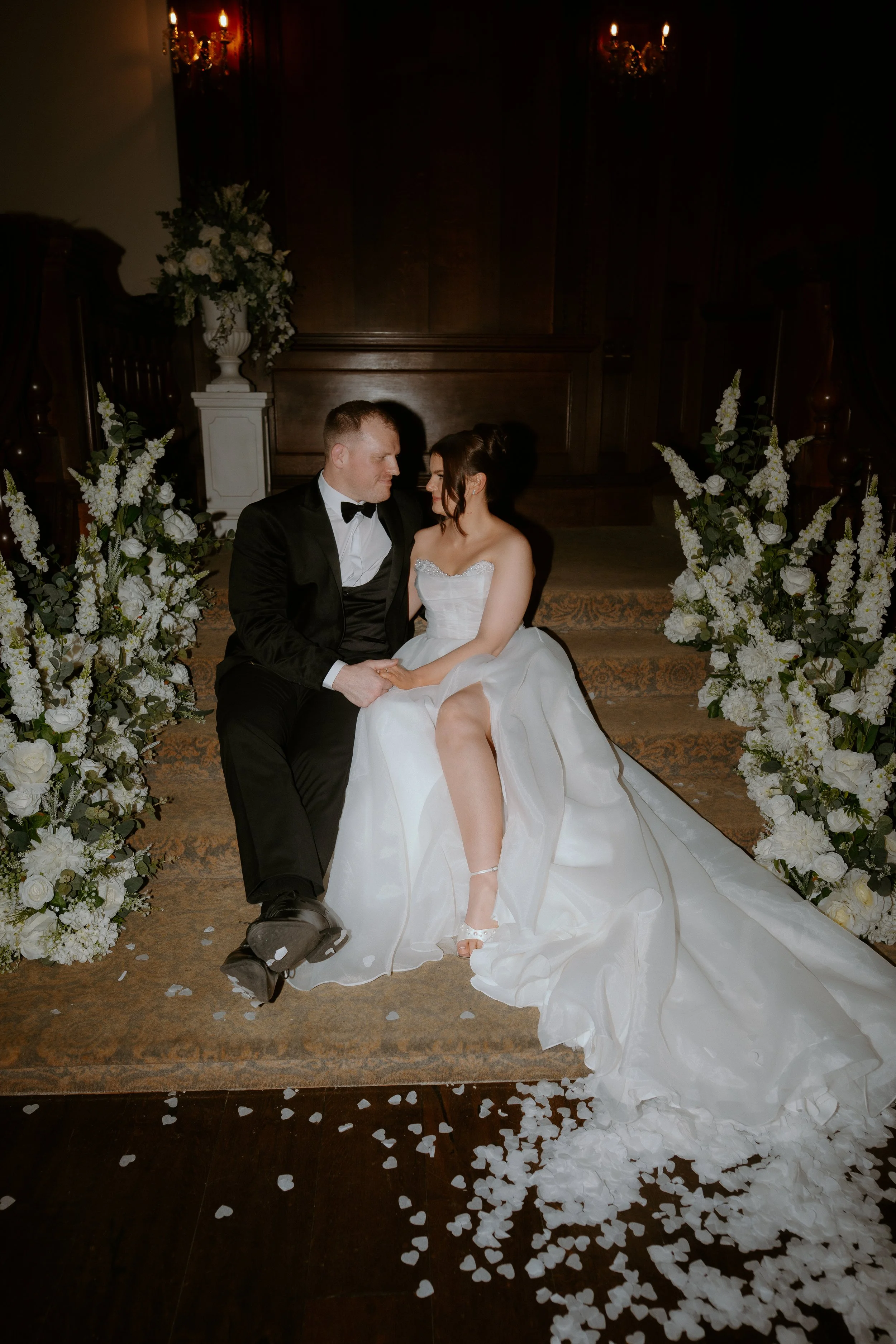 A bride in a white wedding gown and a groom in a black tuxedo sitting on the steps of an altar, holding hands and gazing at each other, surrounded by floral arrangements and white rose petals on the floor.