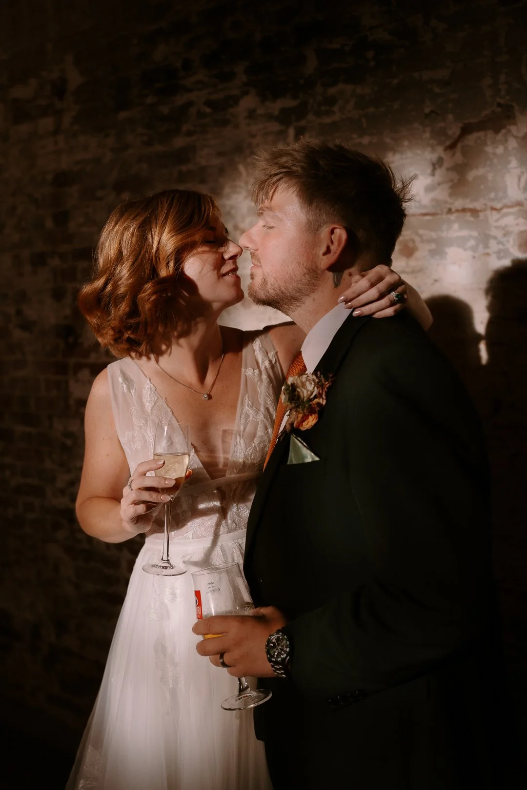 A bride and groom close together, about to kiss, holding drinks at a wedding reception in front of a brick wall.