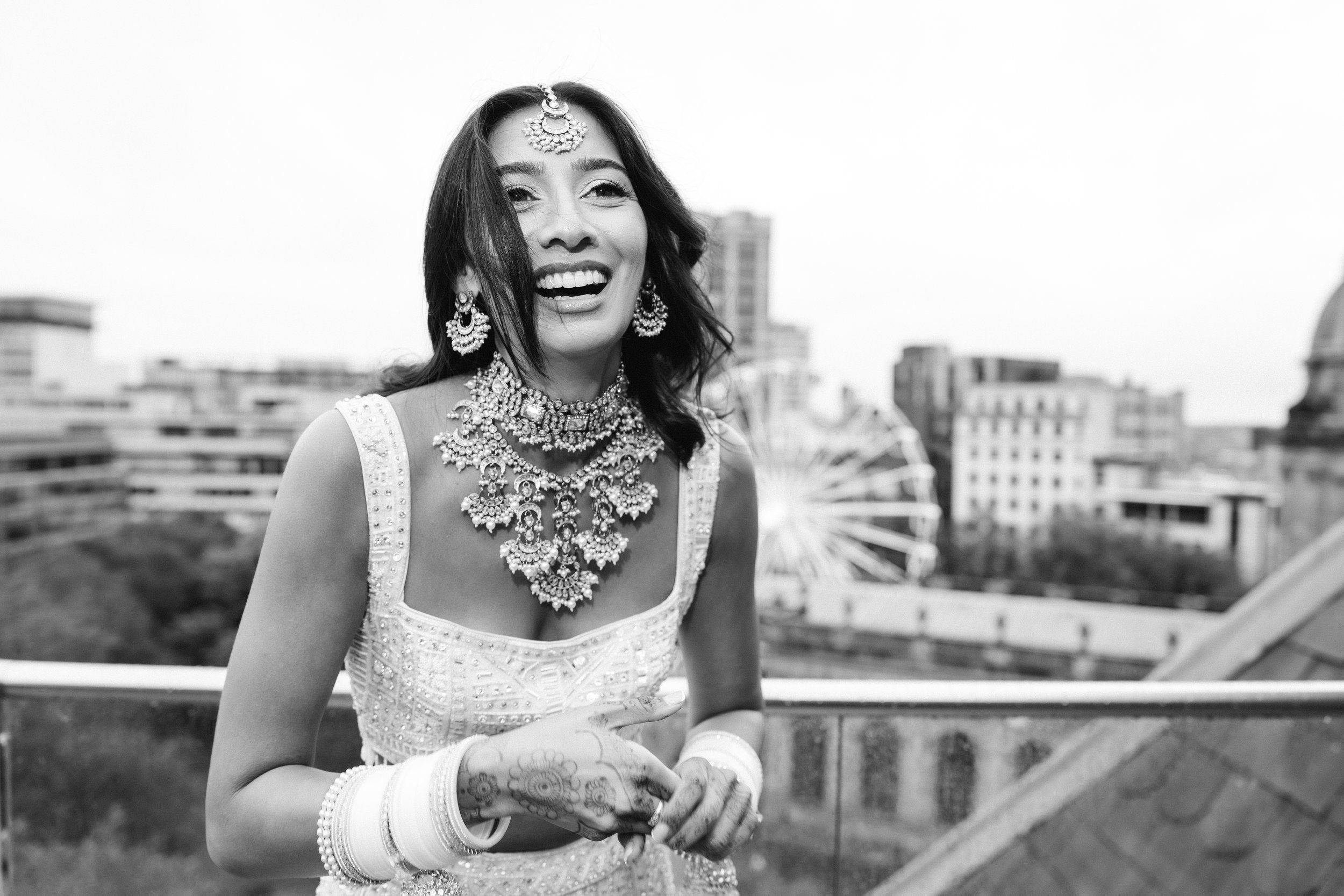 A woman wearing traditional Indian jewelry and attire, smiling and holding her hands together, standing outdoors on a rooftop with city buildings in the background.