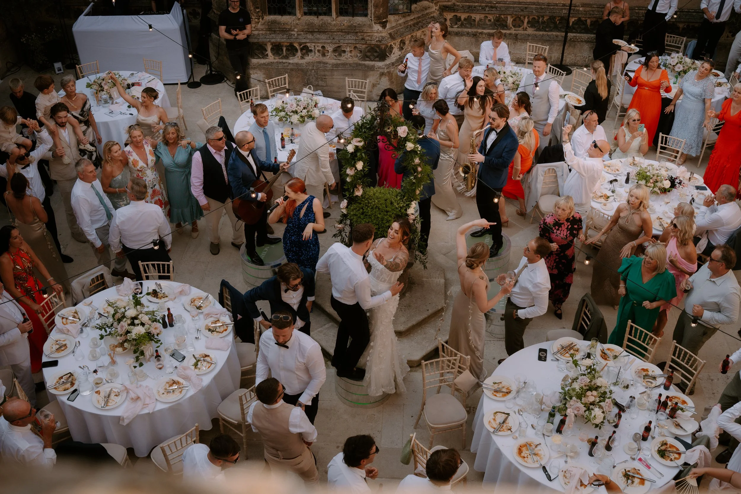 An outdoor wedding reception with round tables decorated with floral centerpieces and placed with dinnerware. Guests are dancing and mingling, with some playing guitars and singing near a floral arch. The setting appears historic with stone architect