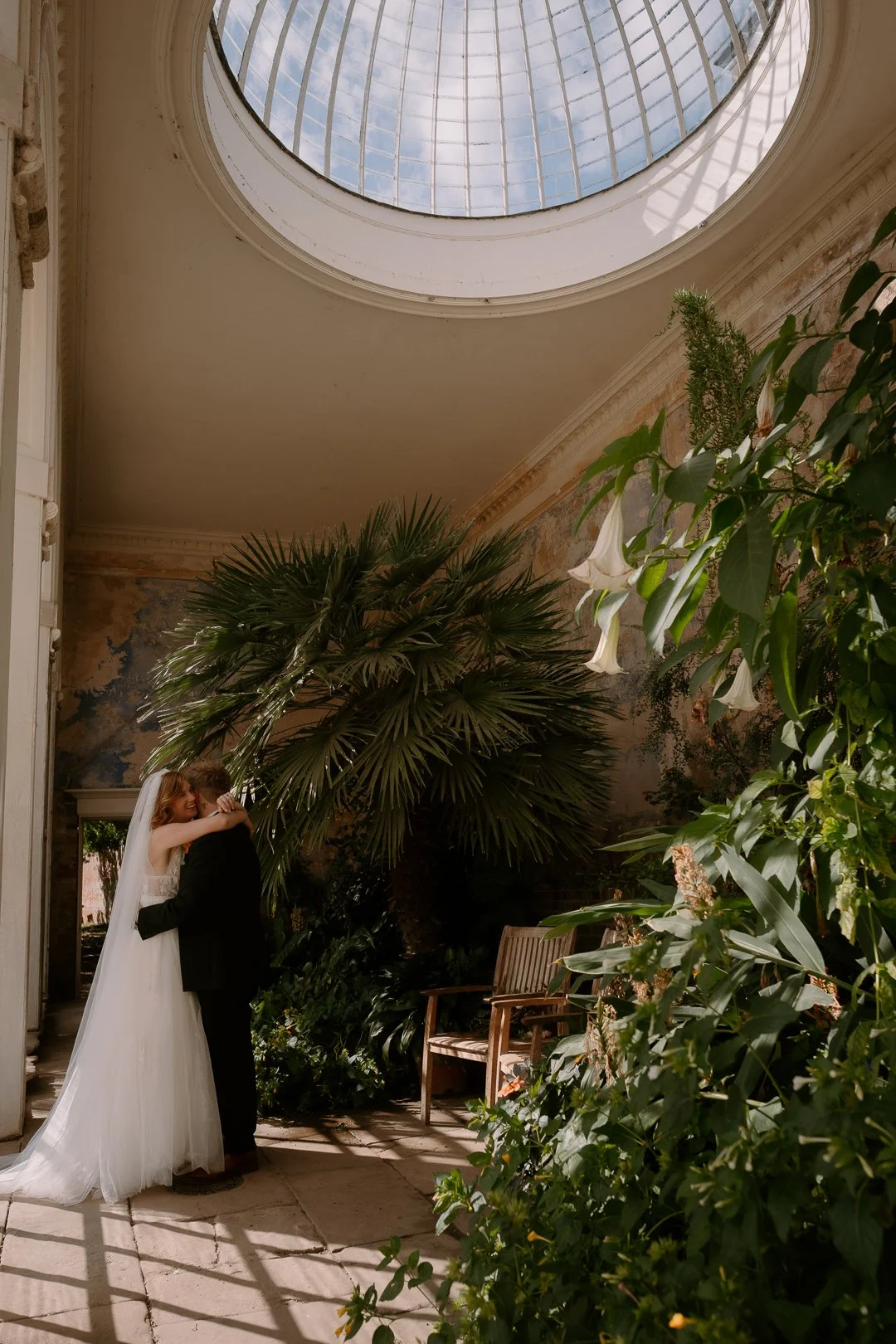 A bride and groom embrace in a lush indoor garden with a glass ceiling overhead, surrounded by green plants and flowers, with a wooden bench nearby.